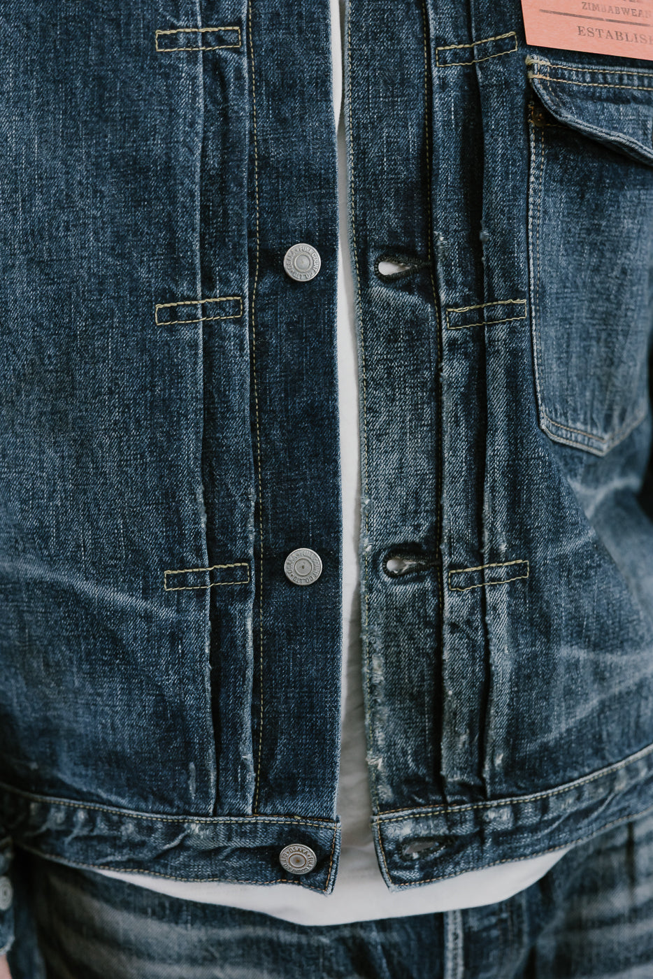 Bearded man wearing Heavily washed indigo denim jacket with a visible full count label on the left chest pocket. Close up of hem, knfie pleats, and donut buttons.