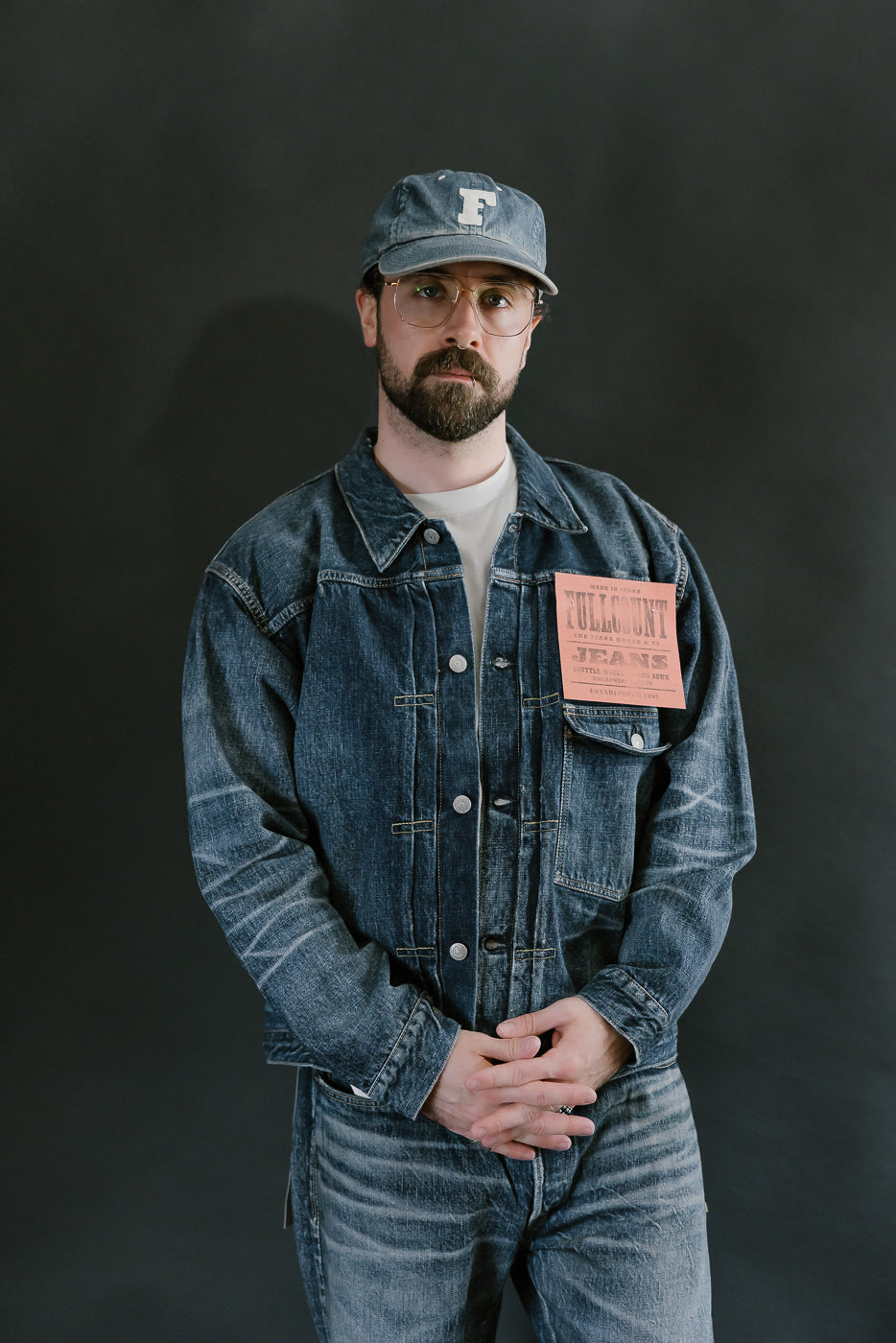 Bearded man wearing Heavily washed indigo denim jacket with a visible full count label on the left chest pocket, heavily washed jeans, pink sneakers, and heavily washed hat with glasses. 3/4 length, front view.