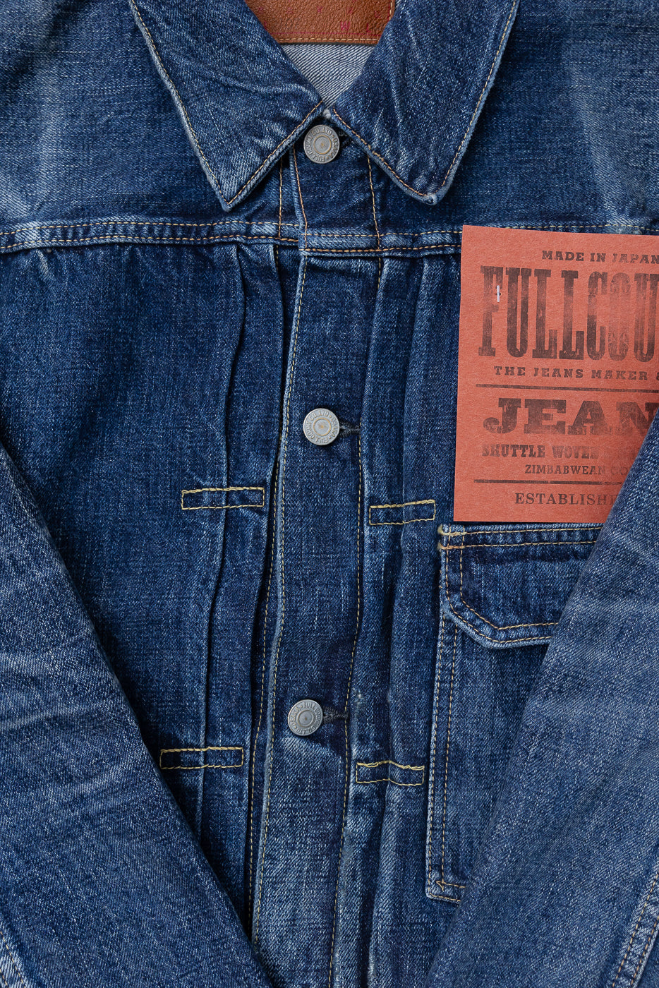 Heavily washed indigo denim jacket with a visible full count label on the left chest pocket on a white background, close up on full count donut buttons and knife pleats.