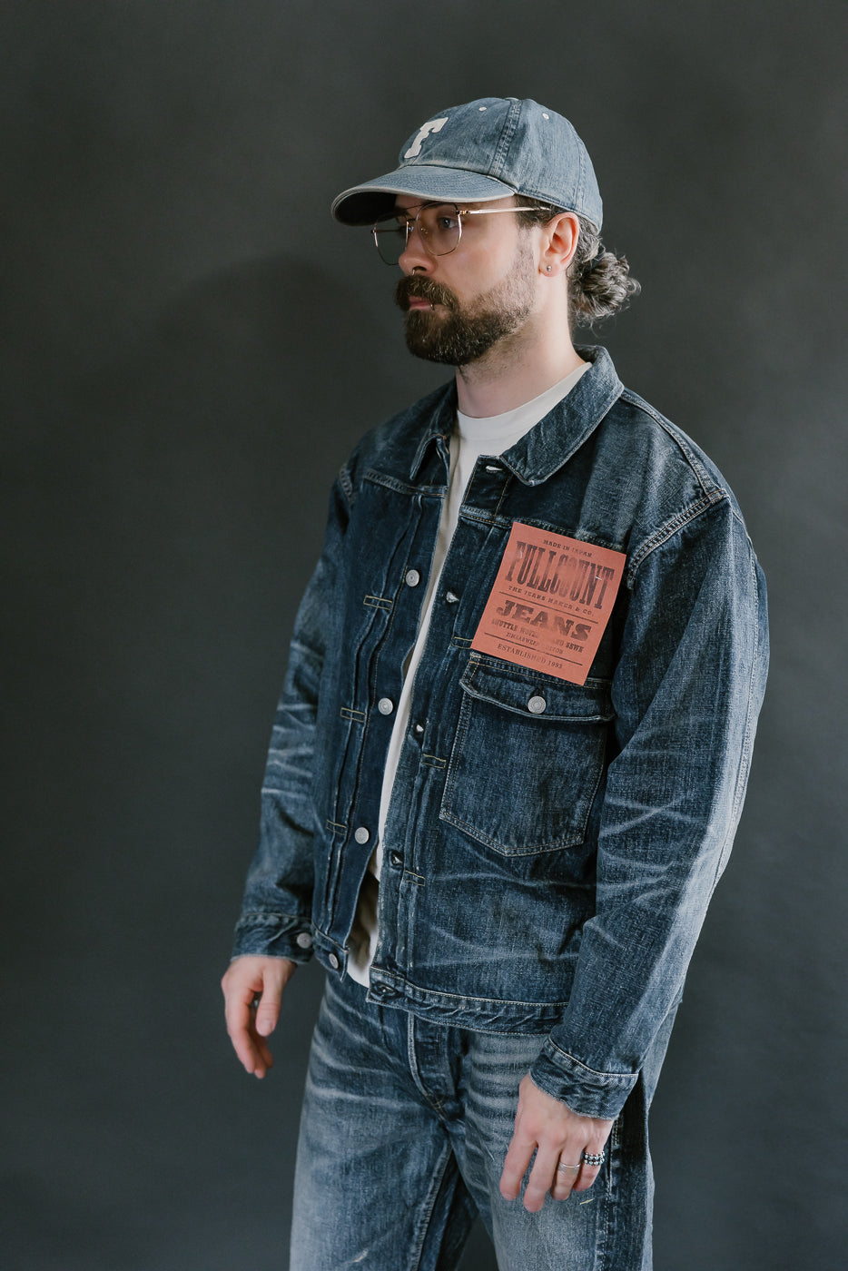 Bearded man wearing Heavily washed indigo denim jacket with a visible full count label on the left chest pocket, heavily washed jeans, pink sneakers, and heavily washed hat with glasses. 3/4 length, side view.