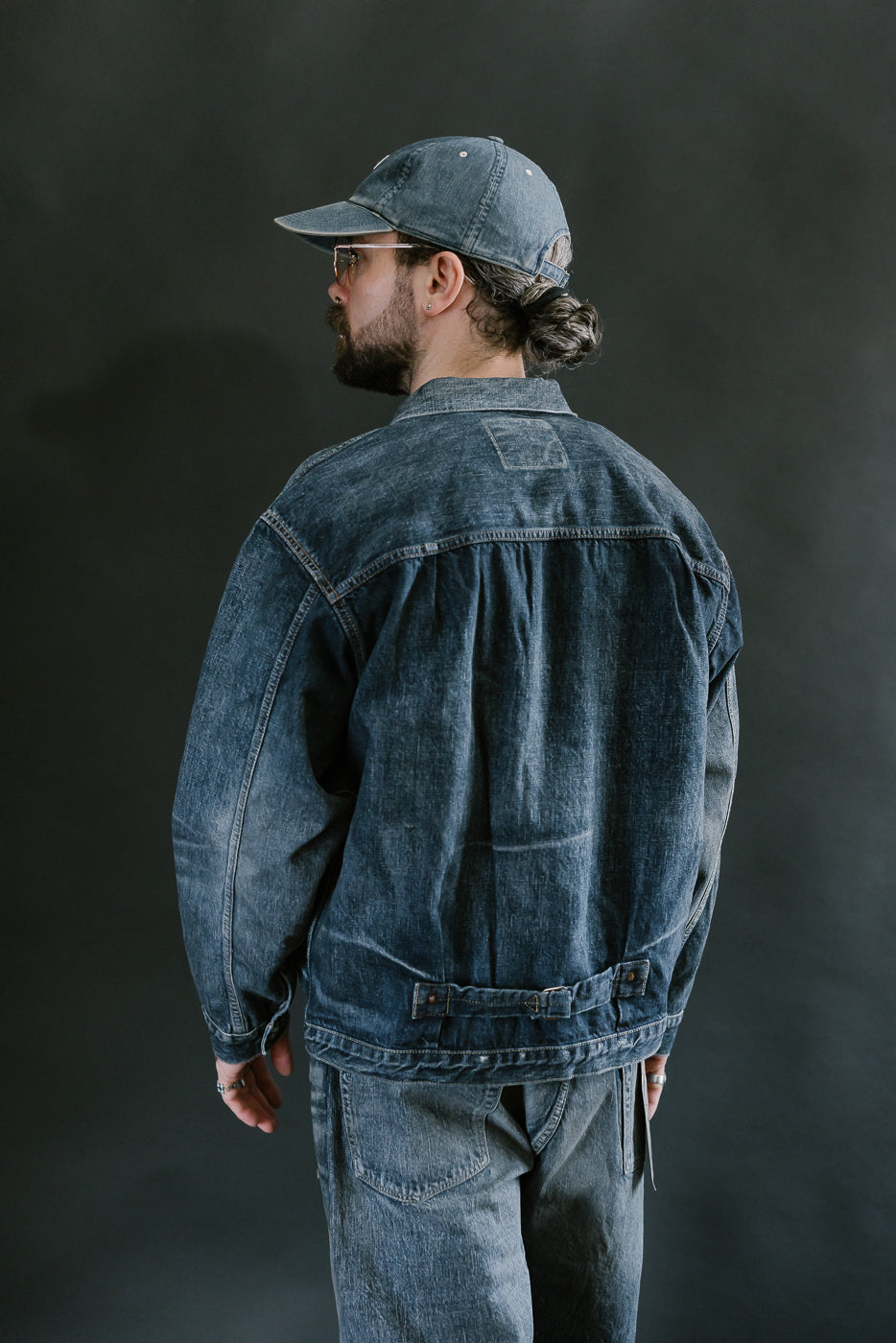 Bearded man wearing Heavily washed indigo denim jacket with a visible full count label on the left chest pocket, heavily washed jeans, and heavily washed hat with glasses. 3/4 length, back view.