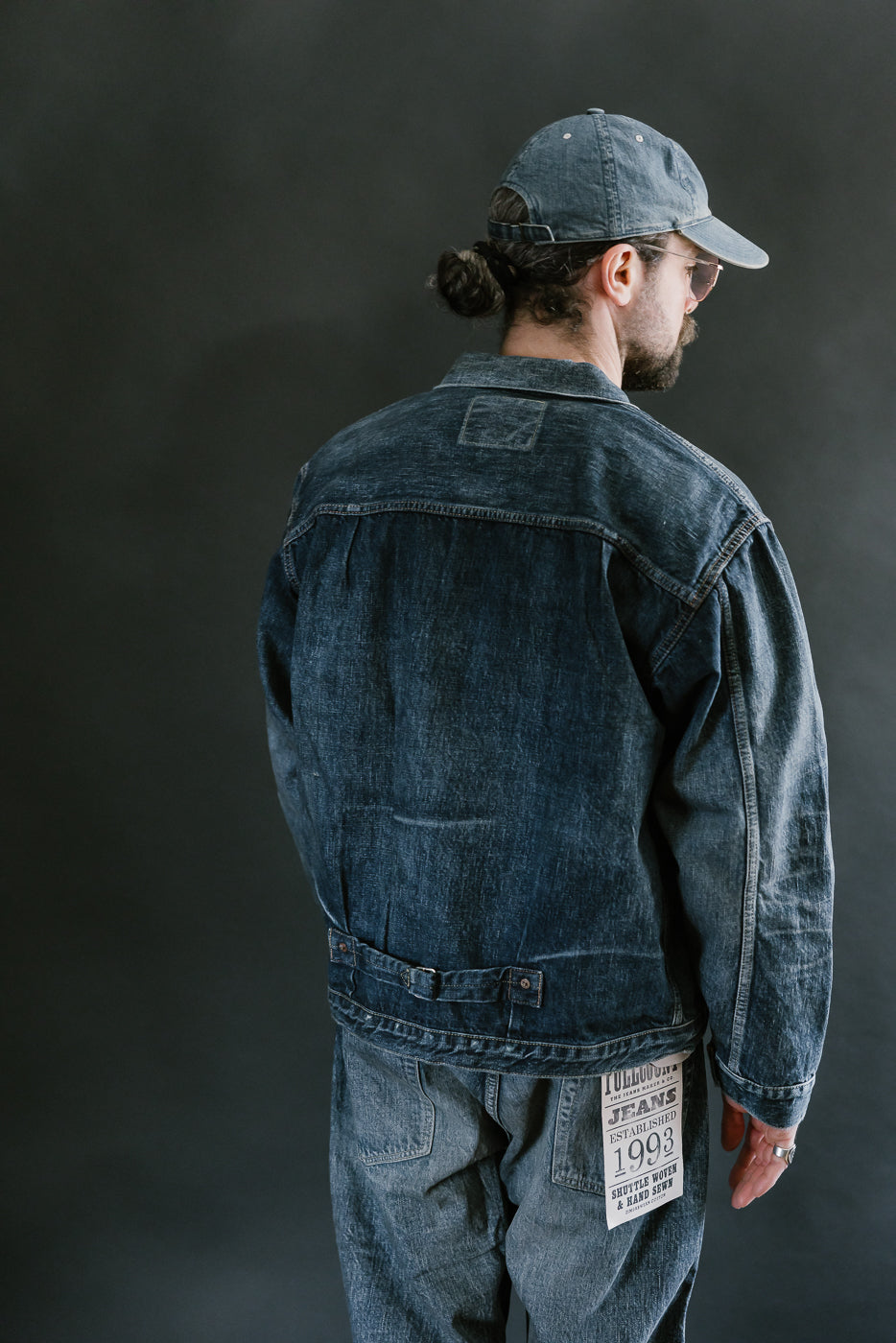 Bearded man wearing Heavily washed indigo denim jacket with a visible full count label on the left chest pocket, heavily washed jeans, and heavily washed hat with glasses. 3/4 length, back view.