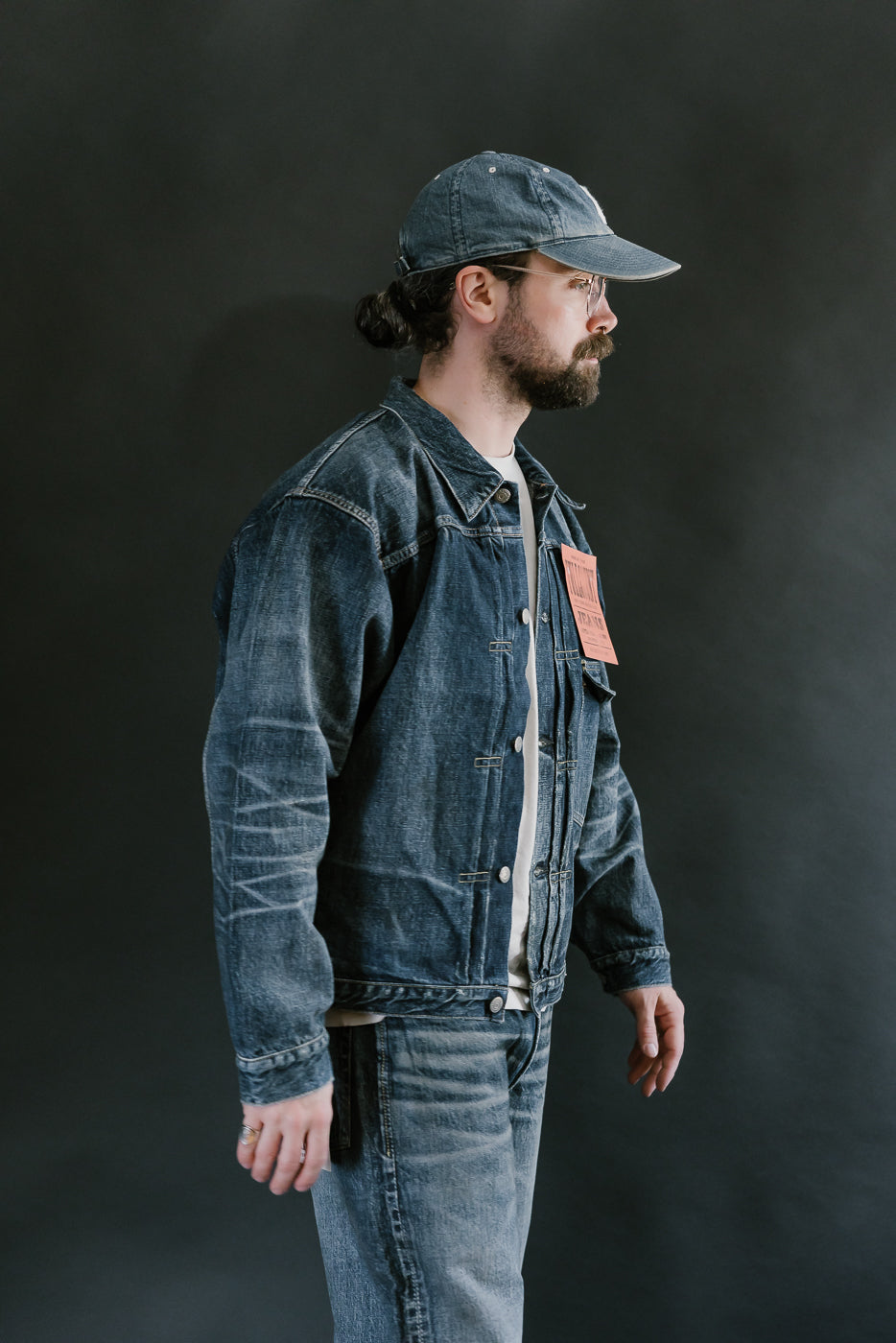 Bearded man wearing Heavily washed indigo denim jacket with a visible full count label on the left chest pocket, heavily washed jeans, and heavily washed hat with glasses. 3/4 length, side view.