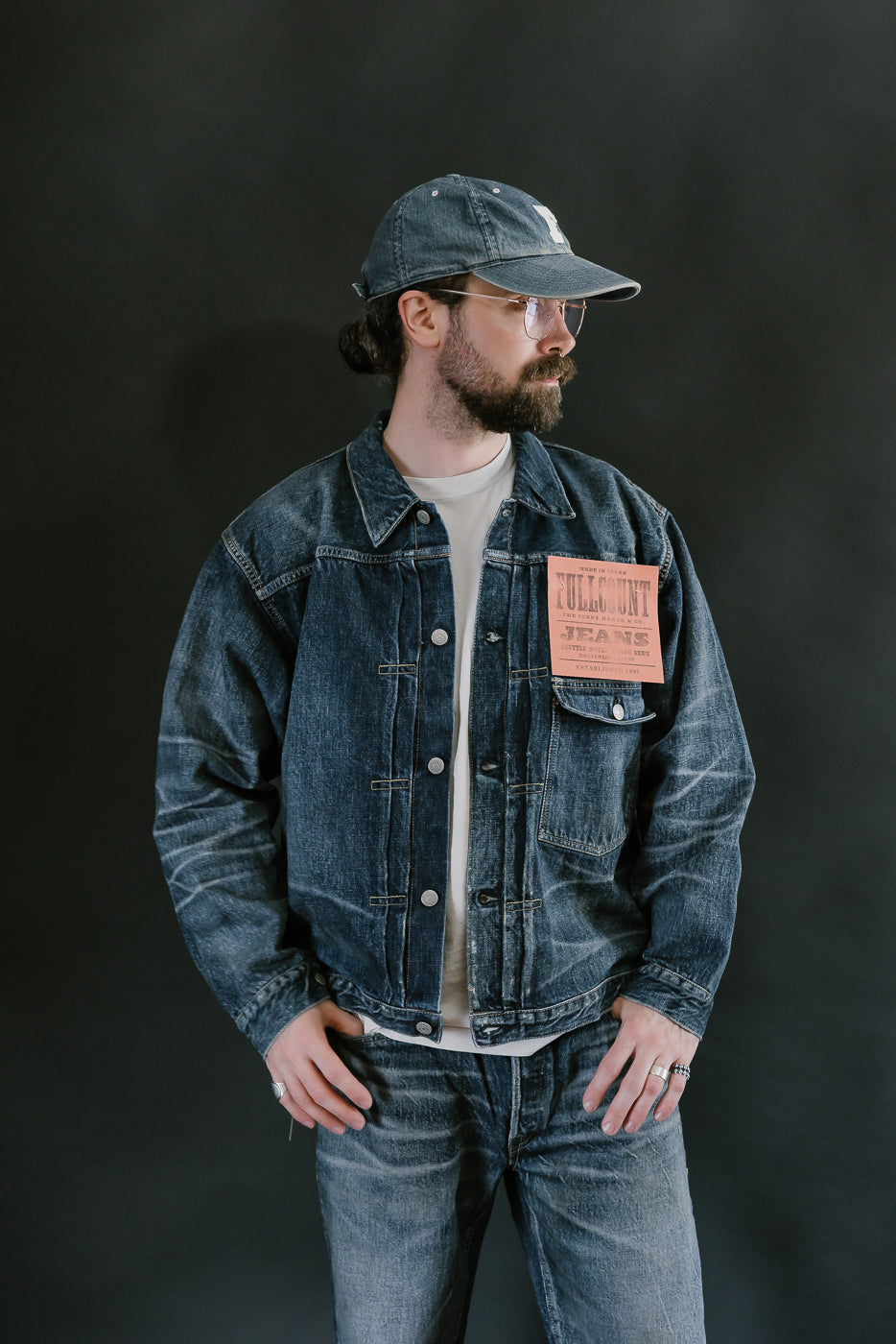 Bearded man wearing Heavily washed indigo denim jacket with a visible full count label on the left chest pocket, heavily washed jeans, and heavily washed hat with glasses. 3/4 length, front view.