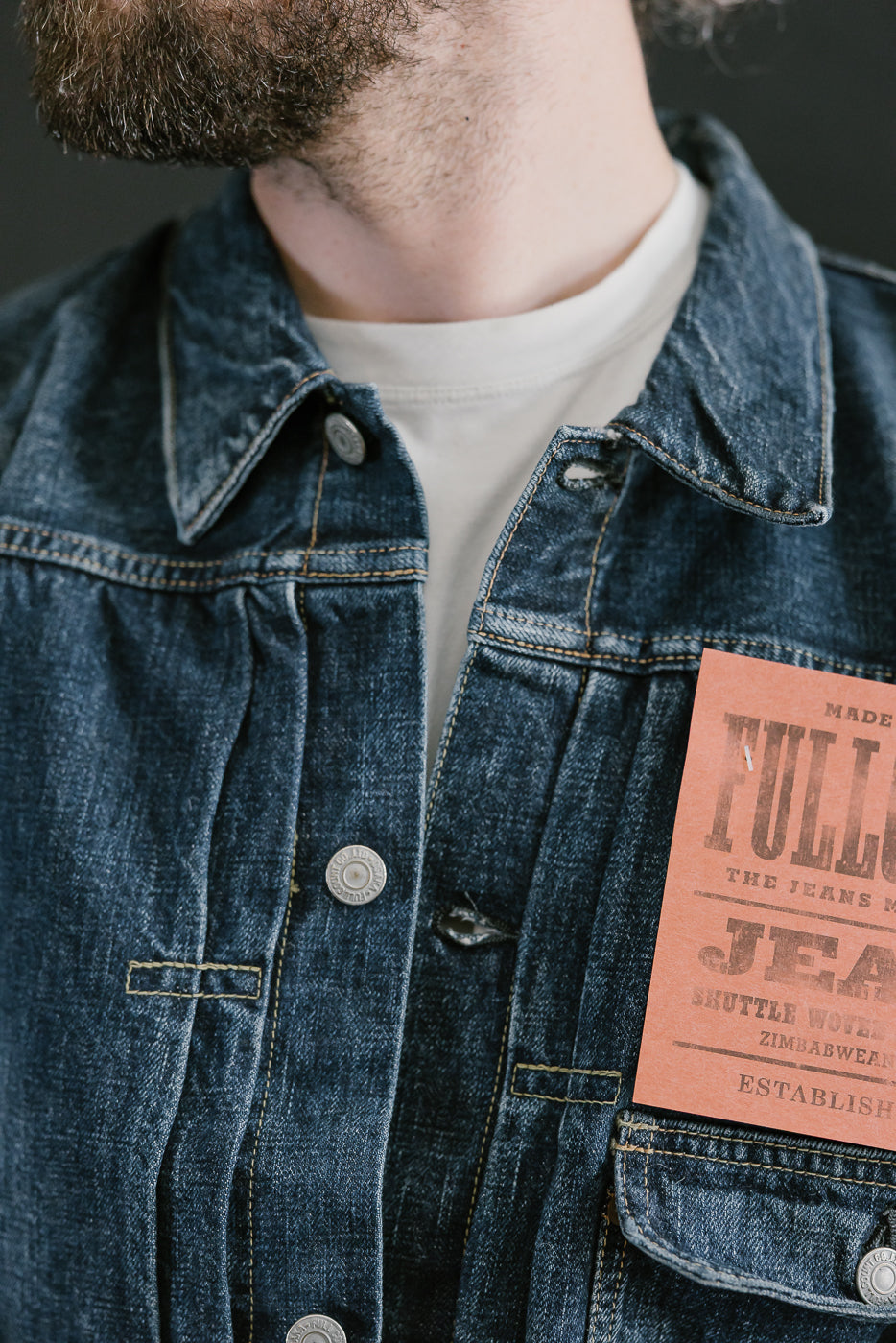 Bearded man wearing Heavily washed indigo denim jacket with a visible full count label on the left chest pocket. Close up of collar and placket.