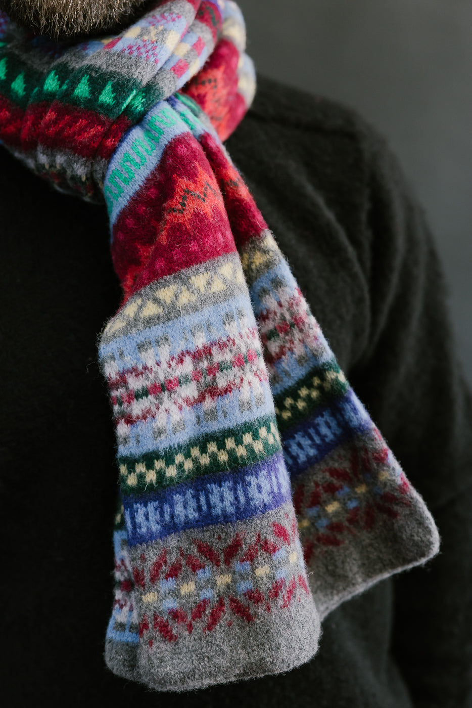multicolored, patterned wool scarf with a heather grey base on a bearded man with a black sweater and chocolate hat, close up