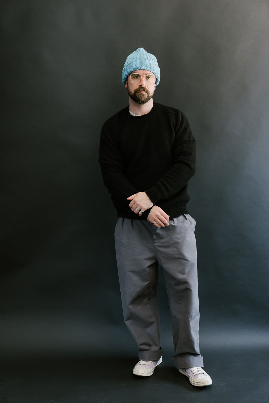 Man wearing a black sweater, gray pants, and a blue beanie against a dark background