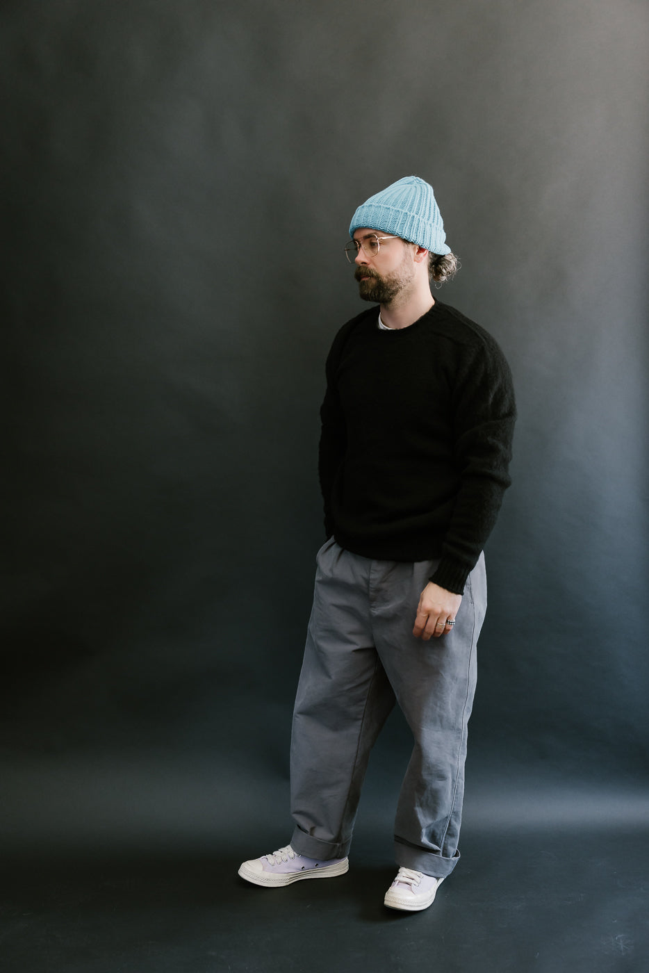 Man wearing a blue beanie, black sweater, gray pants, and white shoes against a dark background