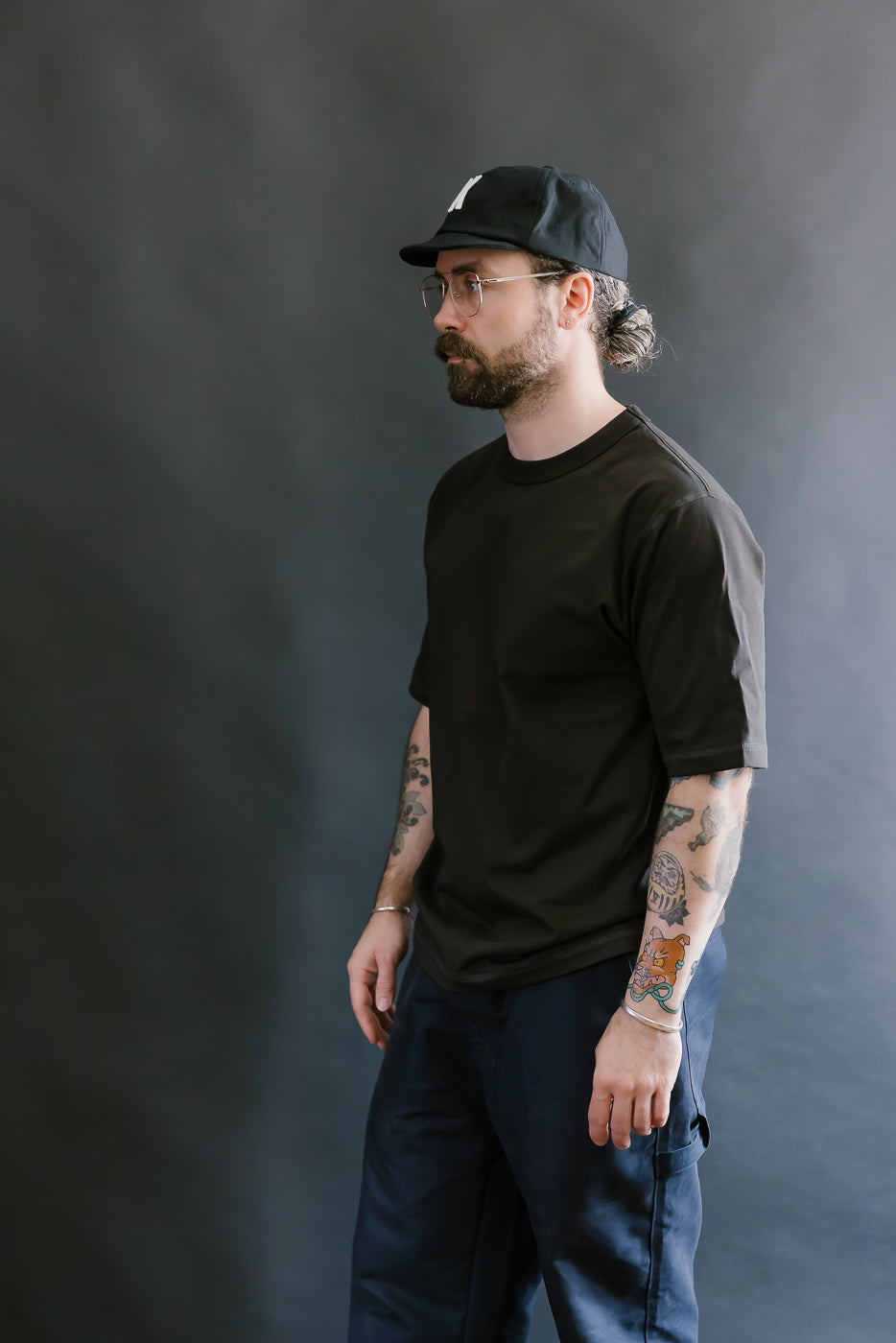 Man wearing a black cap, black t-shirt, and dark pants against a gray background