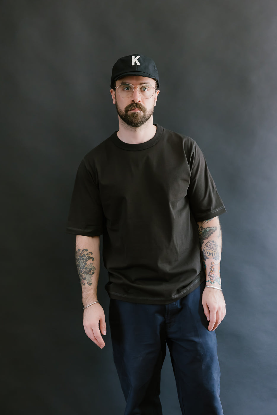 Man wearing a black t-shirt, dark pants, and a black cap with a 'K' logo against a dark background