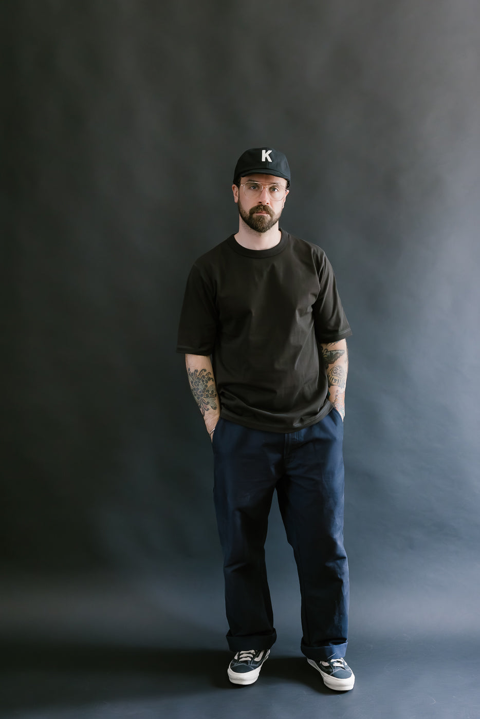 Man wearing a black t-shirt, dark pants, and a black cap with a logo on a gray background