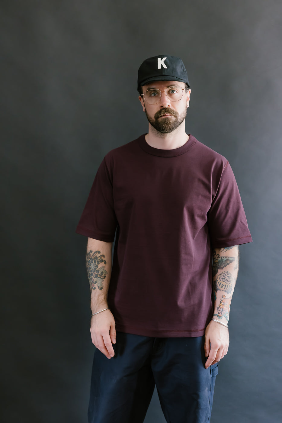 Man wearing a maroon t-shirt and black cap with a 'K' logo against a dark background