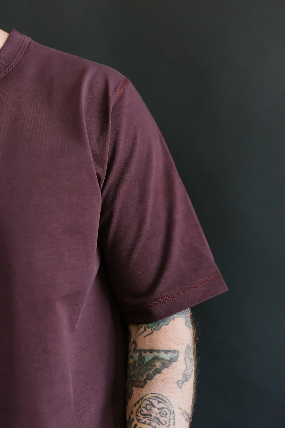Maroon t-shirt worn by a person with tattoos on a black background