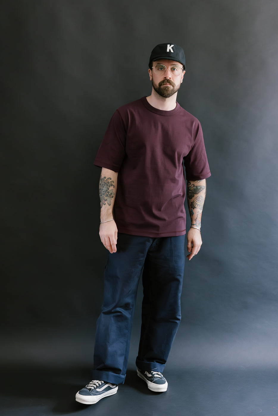 Man wearing a maroon t-shirt, dark pants, and a black cap with a logo on a gray background