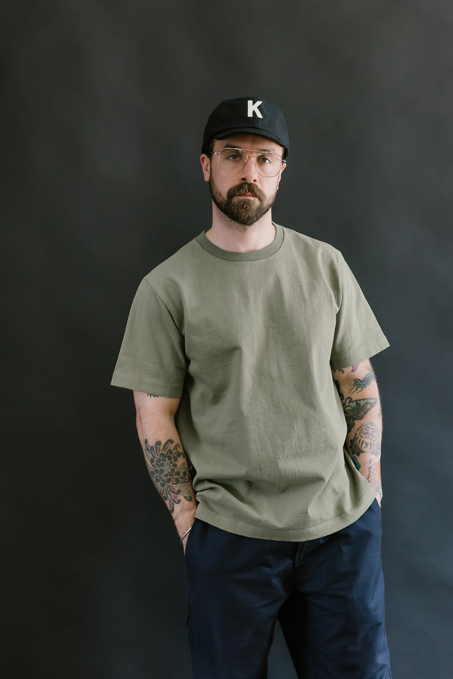 Man wearing a green t-shirt and black cap with a 'K' logo against a dark background