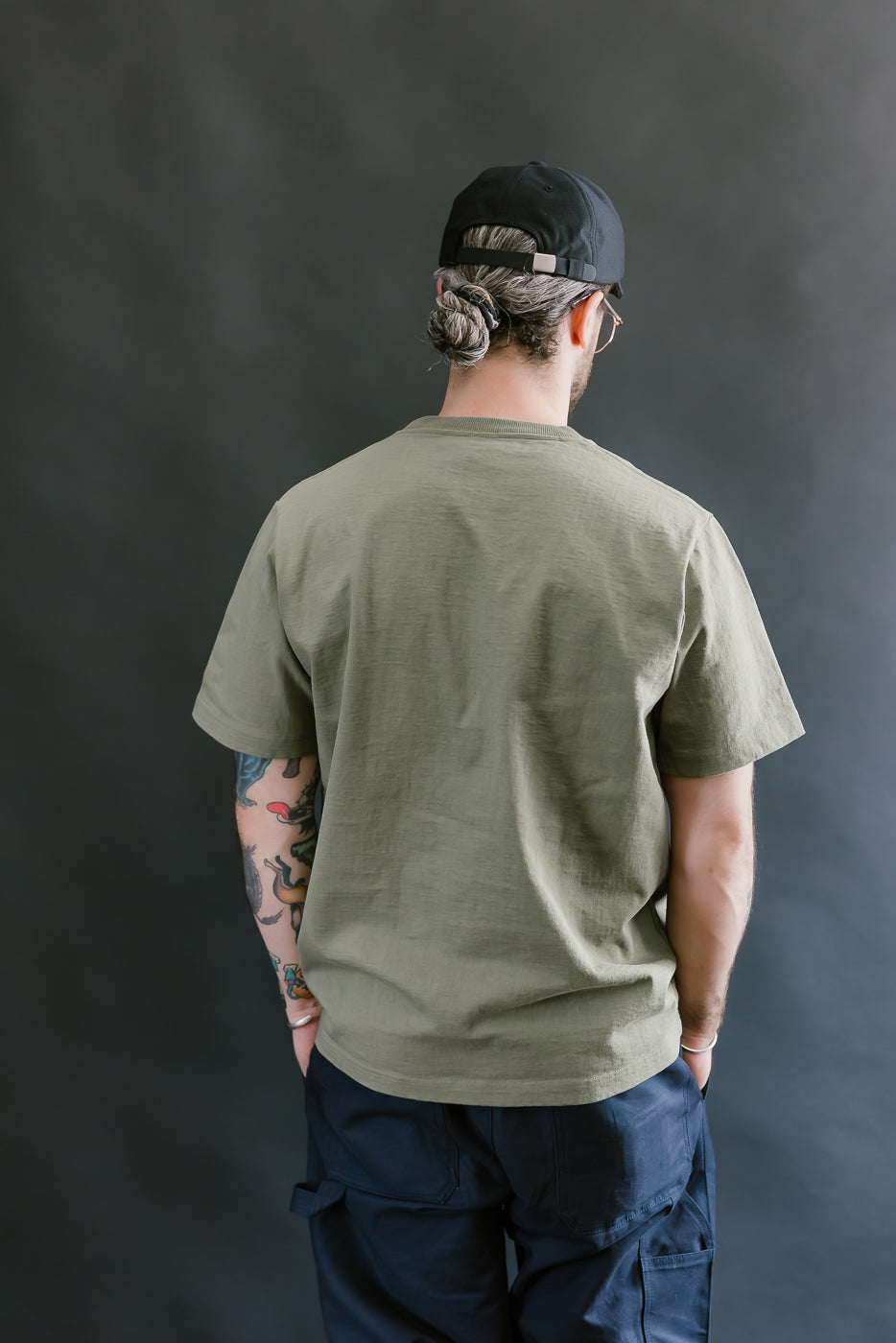 Person wearing an olive green t-shirt and dark pants against a plain background