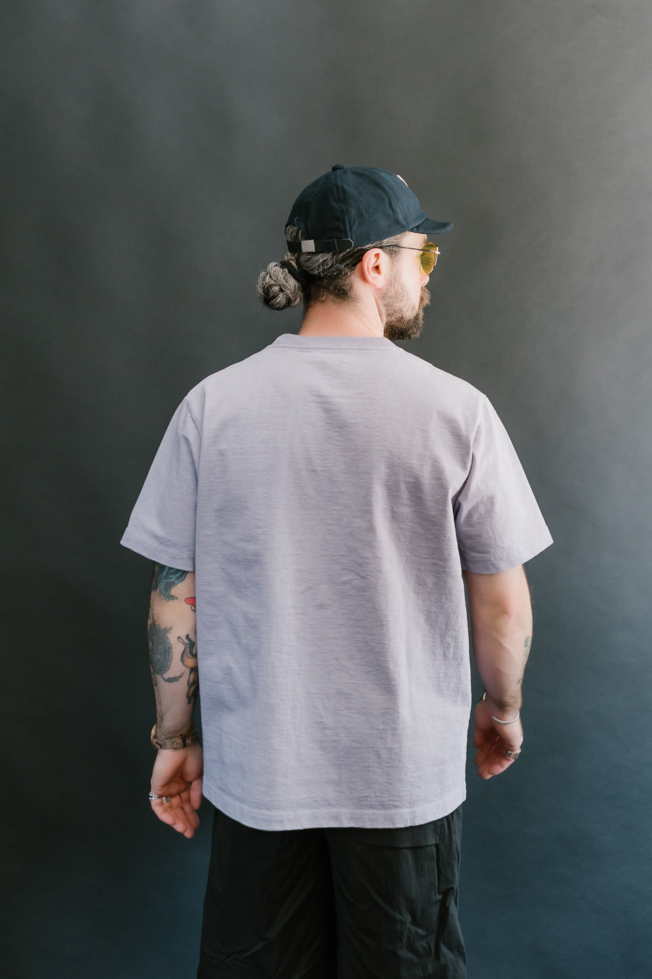 3/4 back view of a man wearing a light purple t-shirt, black cap, and sunglasses against a dark background