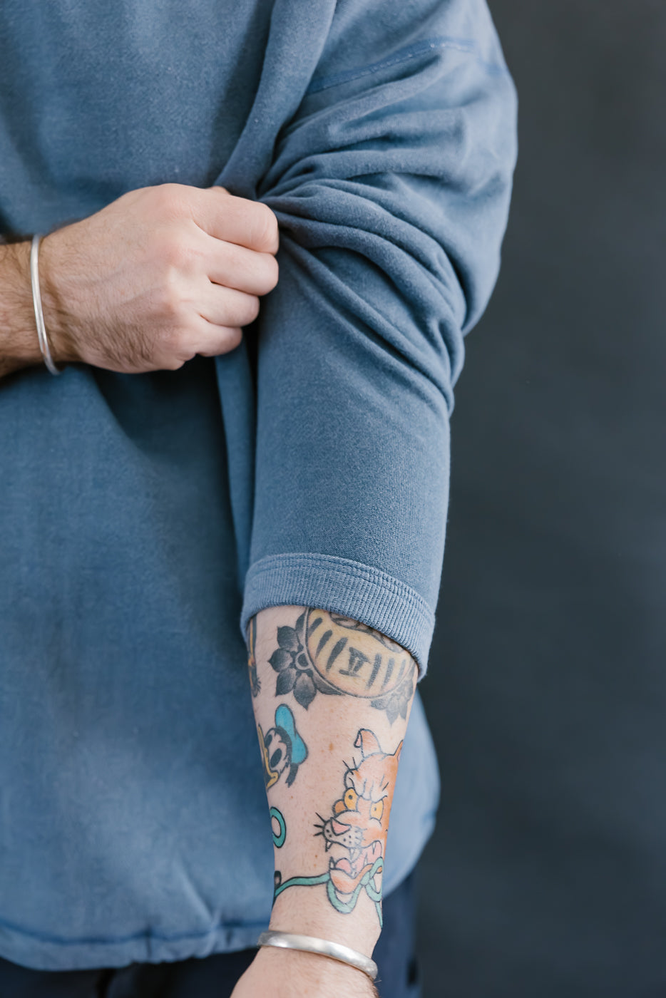 Person wearing a blue sweater with a tattoo on their arm against a dark background