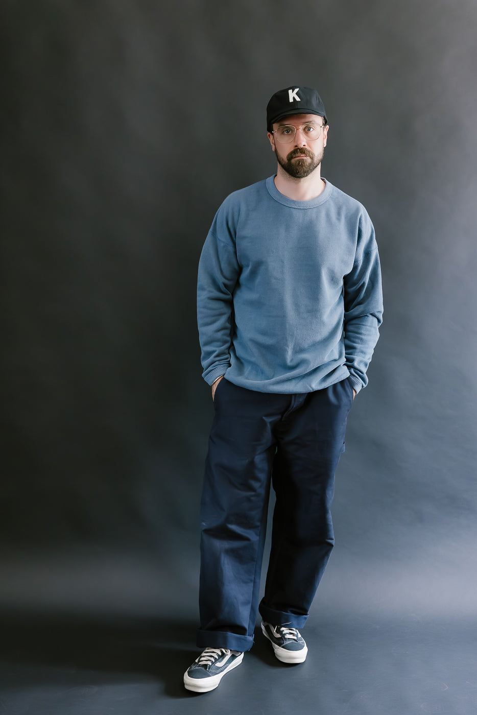 Man wearing a blue sweatshirt, black pants, and a black cap with a 'K' logo against a dark background