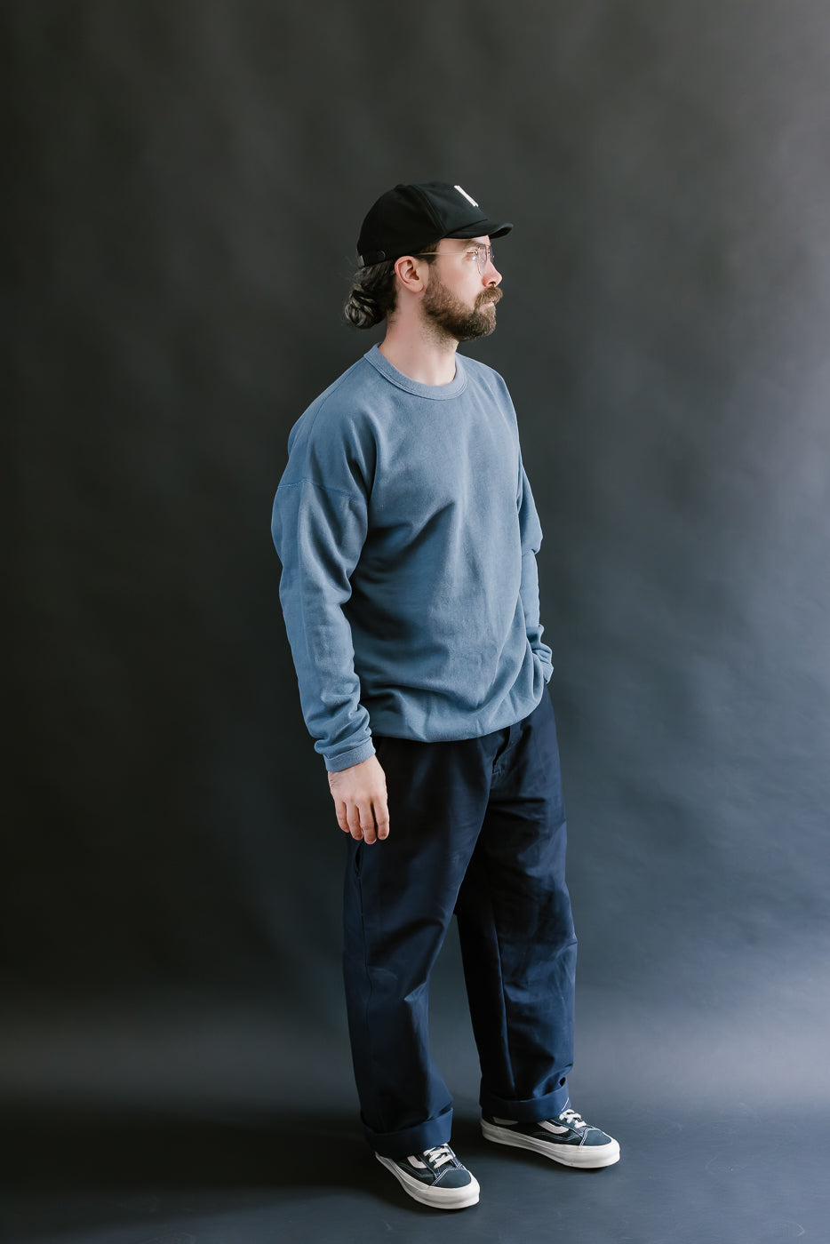 Man wearing a blue sweater, black cap, and dark pants against a gray background