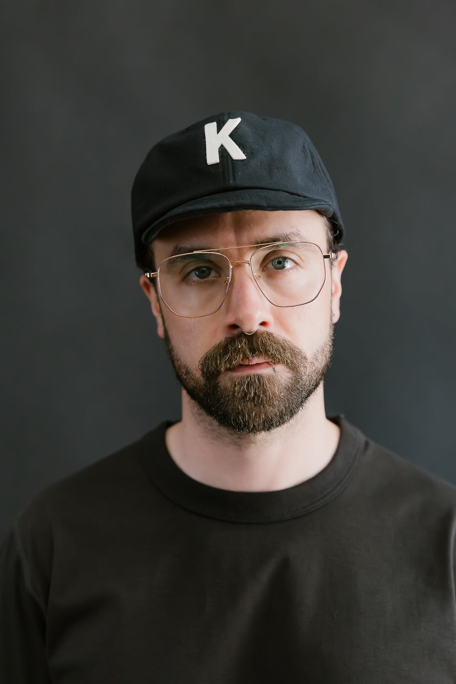 Man wearing a black cap with a white 'K' logo and glasses against a dark background
