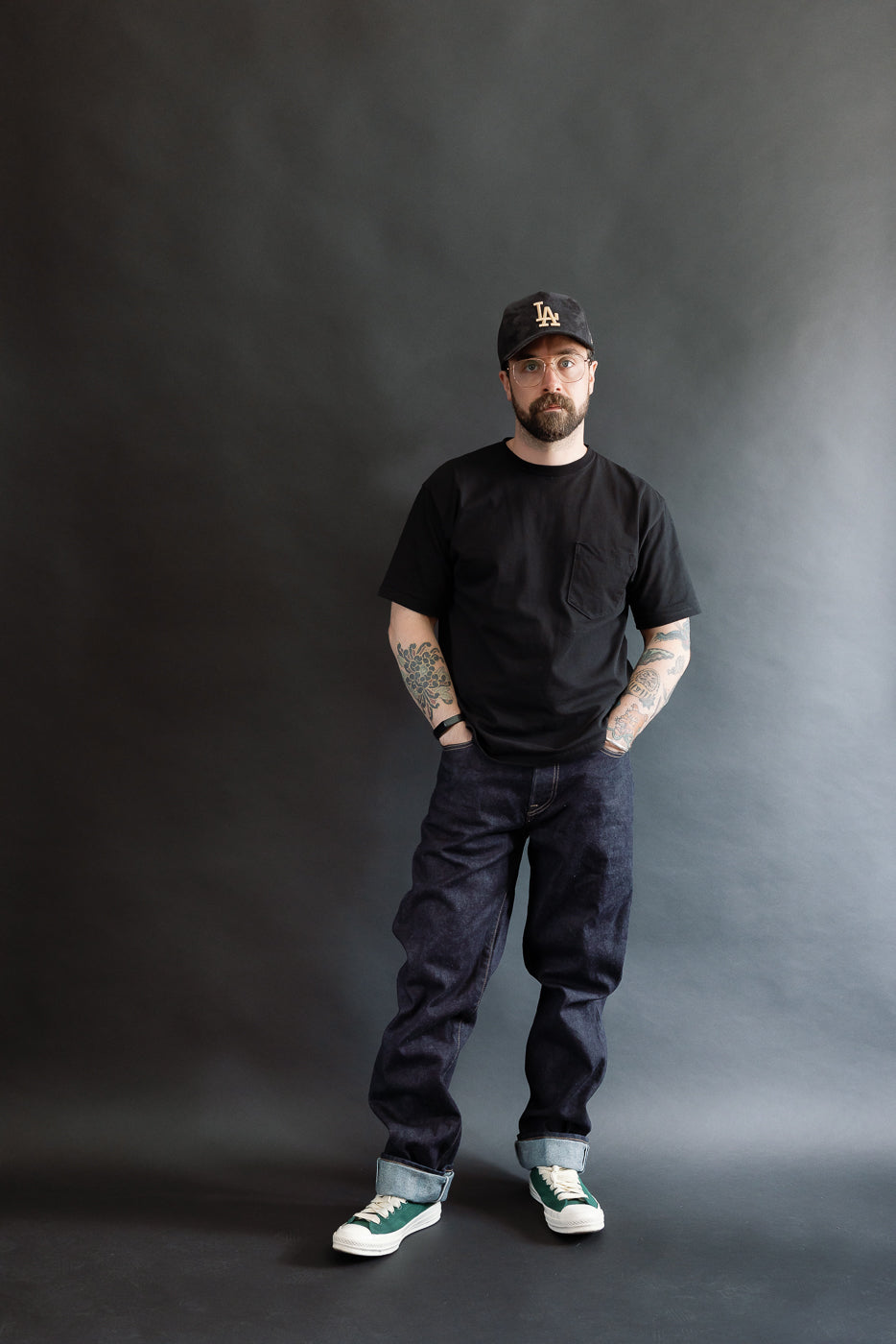 Man wearing a black t-shirt, dark RGT 15oz rinsed selvedge jeans, and a cap against a gray background, front view