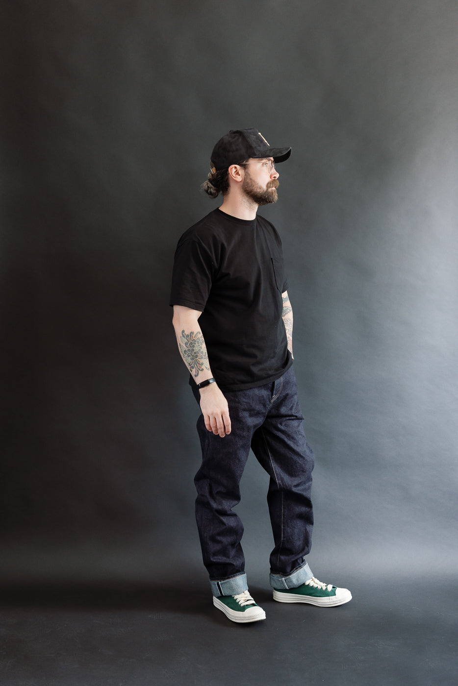Man wearing a black t-shirt, dark RGT 15oz rinsed selvedge jeans, and a cap against a gray background, side view