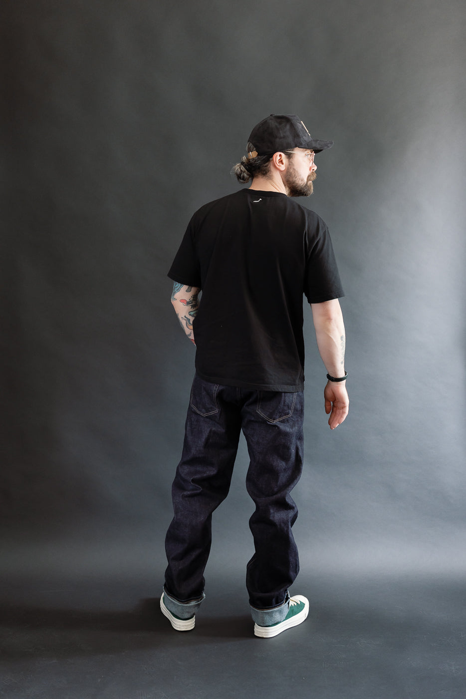 Man wearing a black t-shirt, dark RGT 15oz rinsed selvedge jeans, and a cap against a gray background, back view
