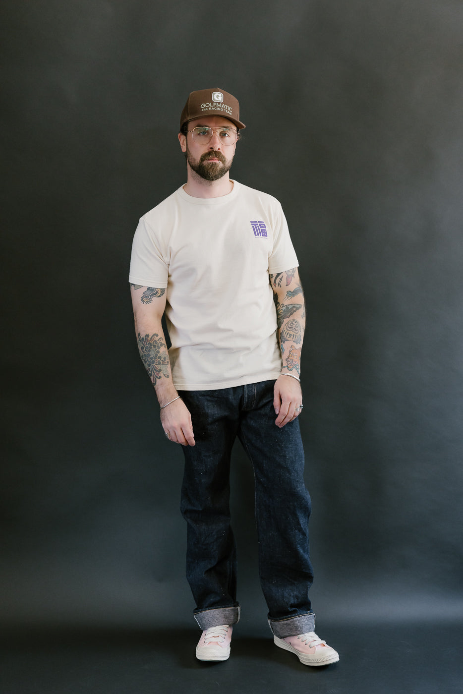 Bearded man wearing S500AX-SAKURA - 18oz Ai-Plus-Sakura Selvedge Jeans with rolled cuffs, styled with a cream T-shirt, baseball cap, and pink sneakers in a studio setting.