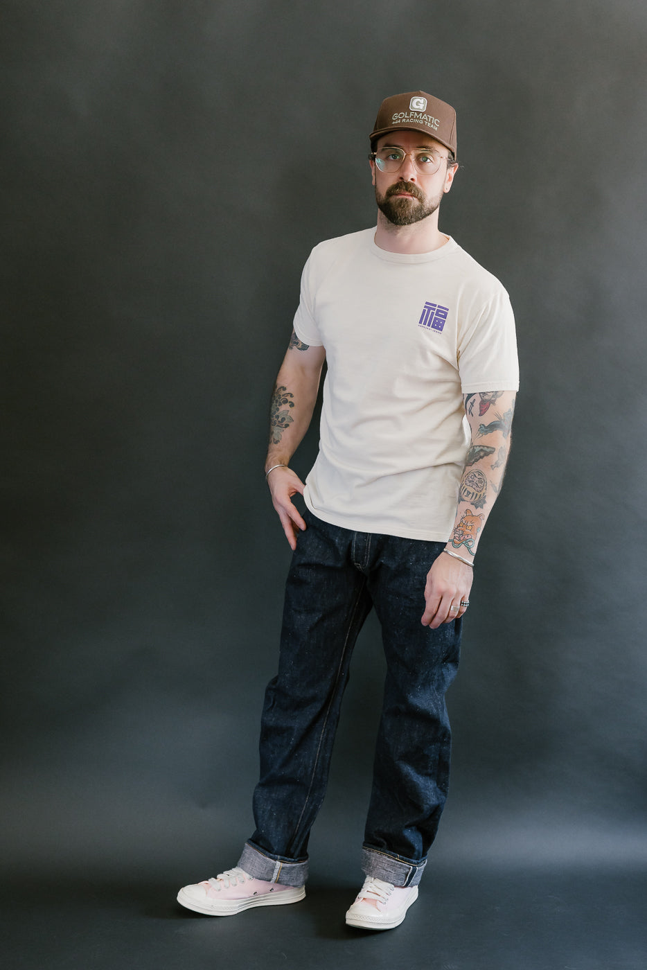 Bearded man wearing Samurai's S500AX-SAKURA jeans, showcasing deep indigo denim with sakura-dyed weft, paired with a beige T-shirt and pink sneakers.