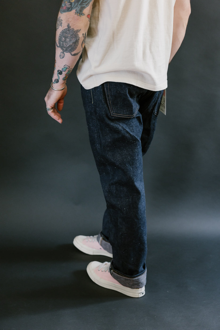 Person wearing Samurai S500AX-SAKURA - 18oz Ai-Plus-Sakura Selvedge Jeans with cuffed ankles, showcasing Japanese indigo denim and sakura-dyed weft, alongside pink and gray canvas sneakers.