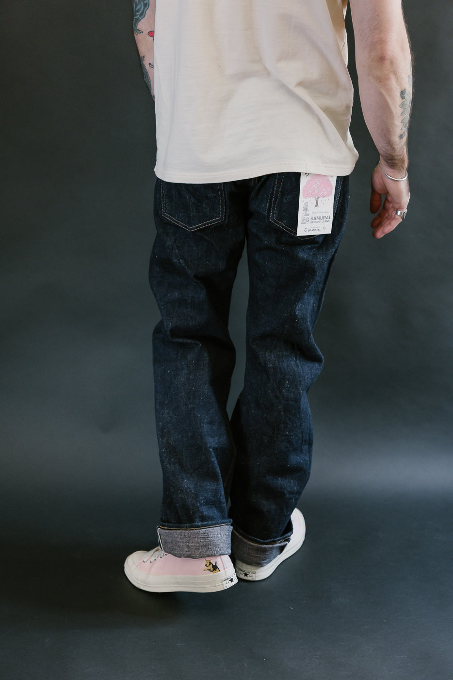 S500AX-SAKURA jeans feature a straight cut, raw indigo denim with sakura-dyed weft, rolled cuffs, and sakura motif details, paired with pink canvas sneakers.