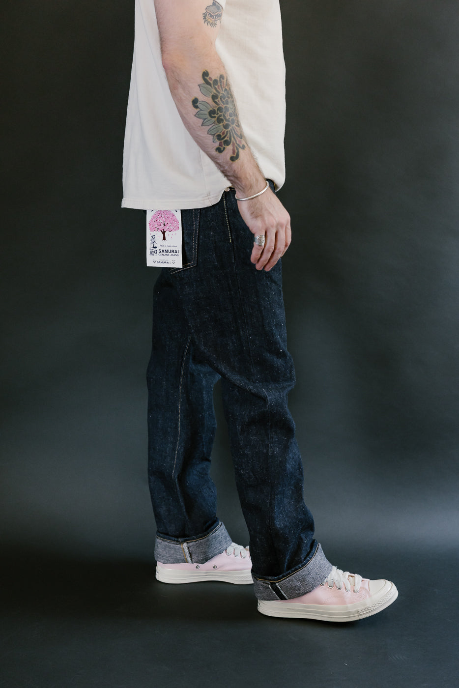 Person wearing Samurai S500AX-SAKURA jeans with cherry-blossom tag, off-white T-shirt, pale pink sneakers, and visible floral tattoo, ring, and bangle.