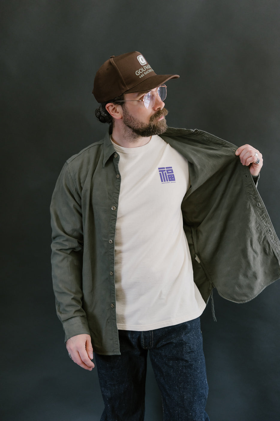 Man wearing SSS25-ATW Tropical Weather Dobby Shirt in olive green, showcasing a stylized graphic on his T-shirt, paired with dark denim jeans.