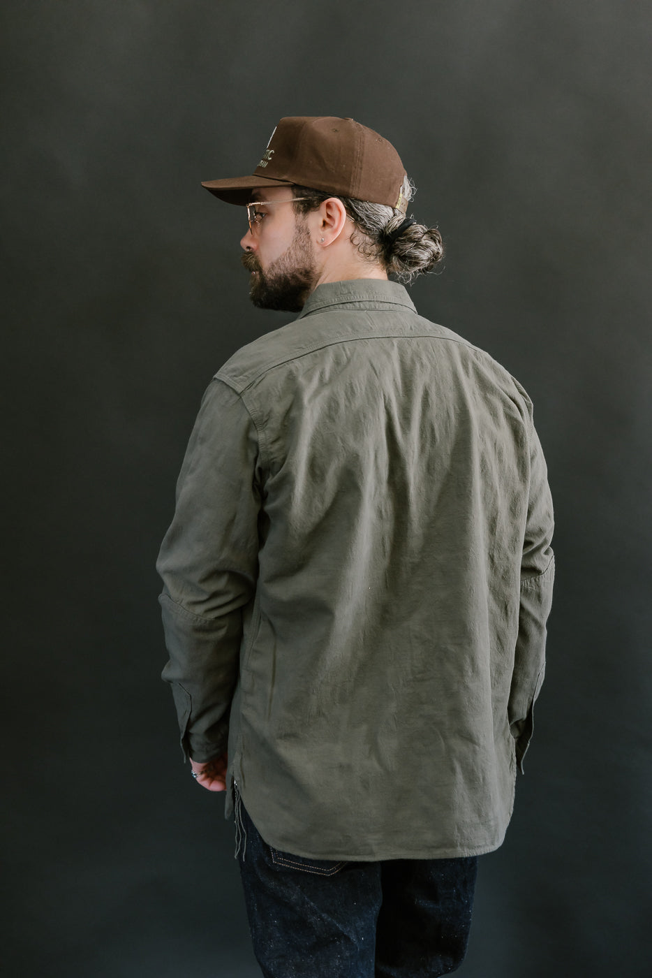 Man in profile wearing SSS25-ATW Tropical Weather Dobby Shirt, olive green, showcasing its classic fit and craftsmanship, paired with jeans and a cap.