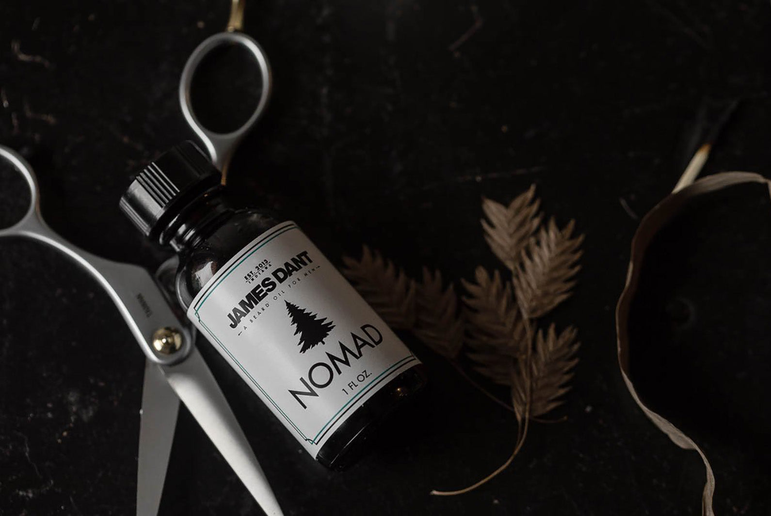 James Dant Nomad beard oil bottle with scissors and leaf on dark surface, Dant contemporary accessories
