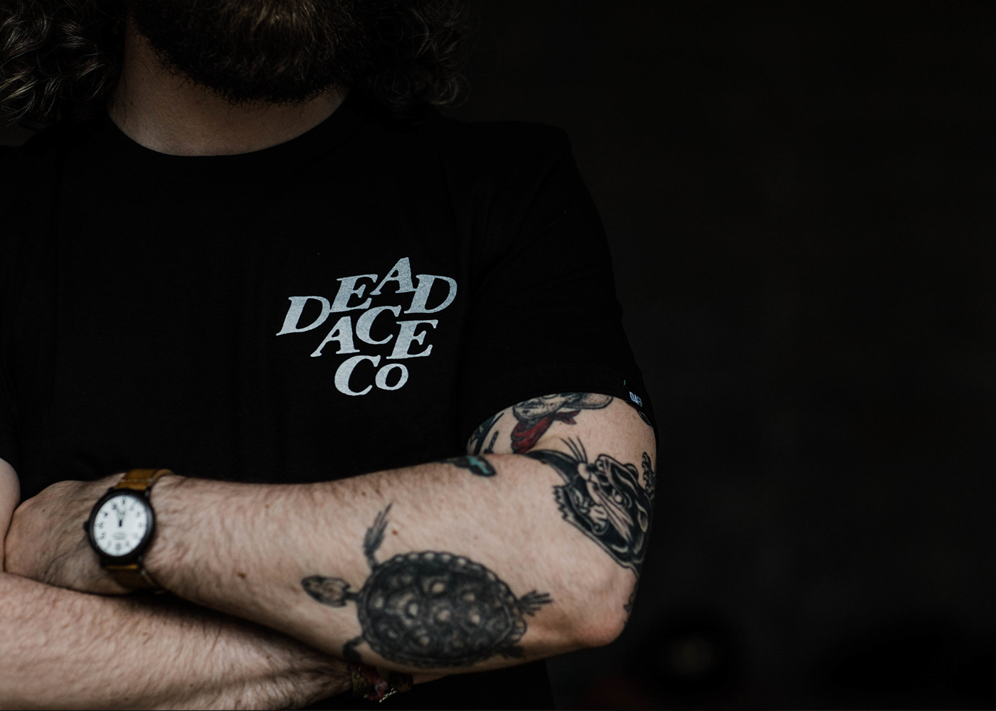 Man in black Dead Ace Co t-shirt with tattoos and watch, contemporary casual apparel