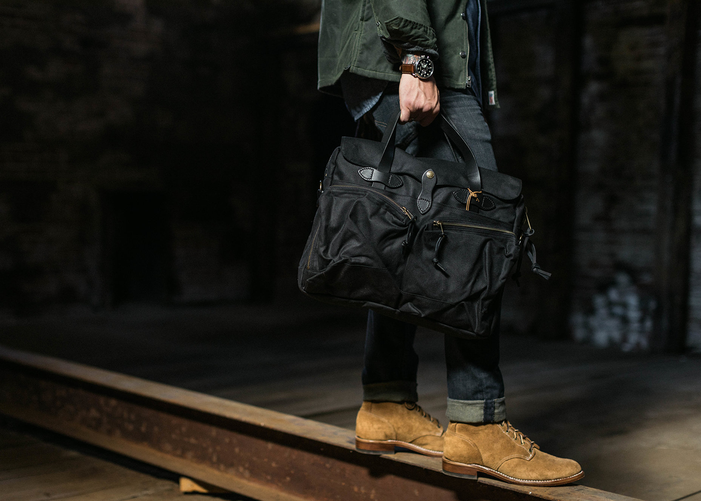 Man holding black duffle bag, wearing suede boots and jeans, in an industrial setting