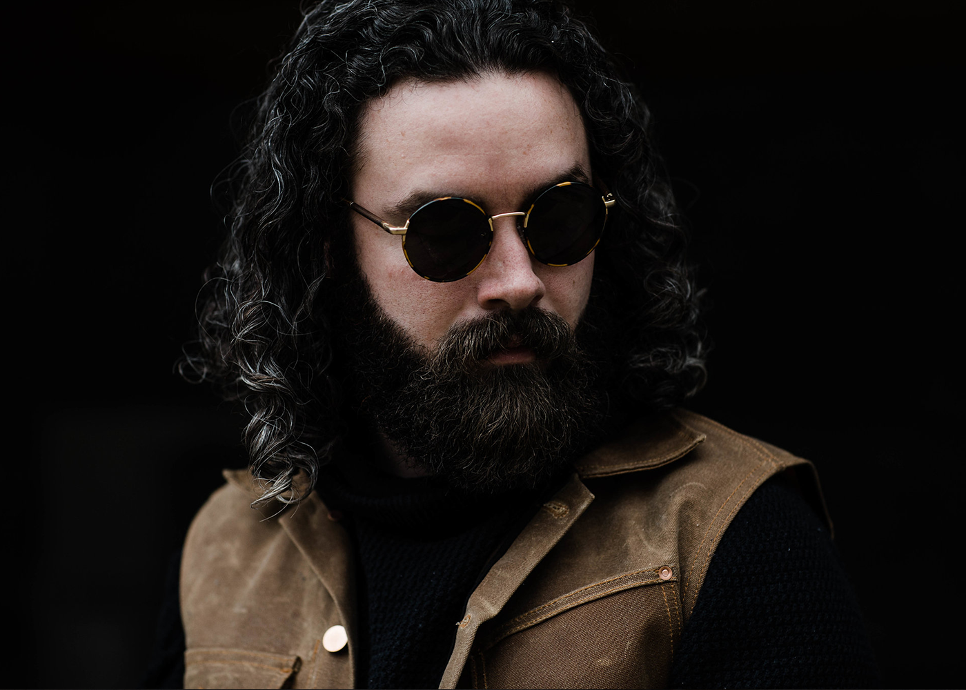 Bearded man with curly hair wearing sunglasses and brown work vest, contemporary casual style