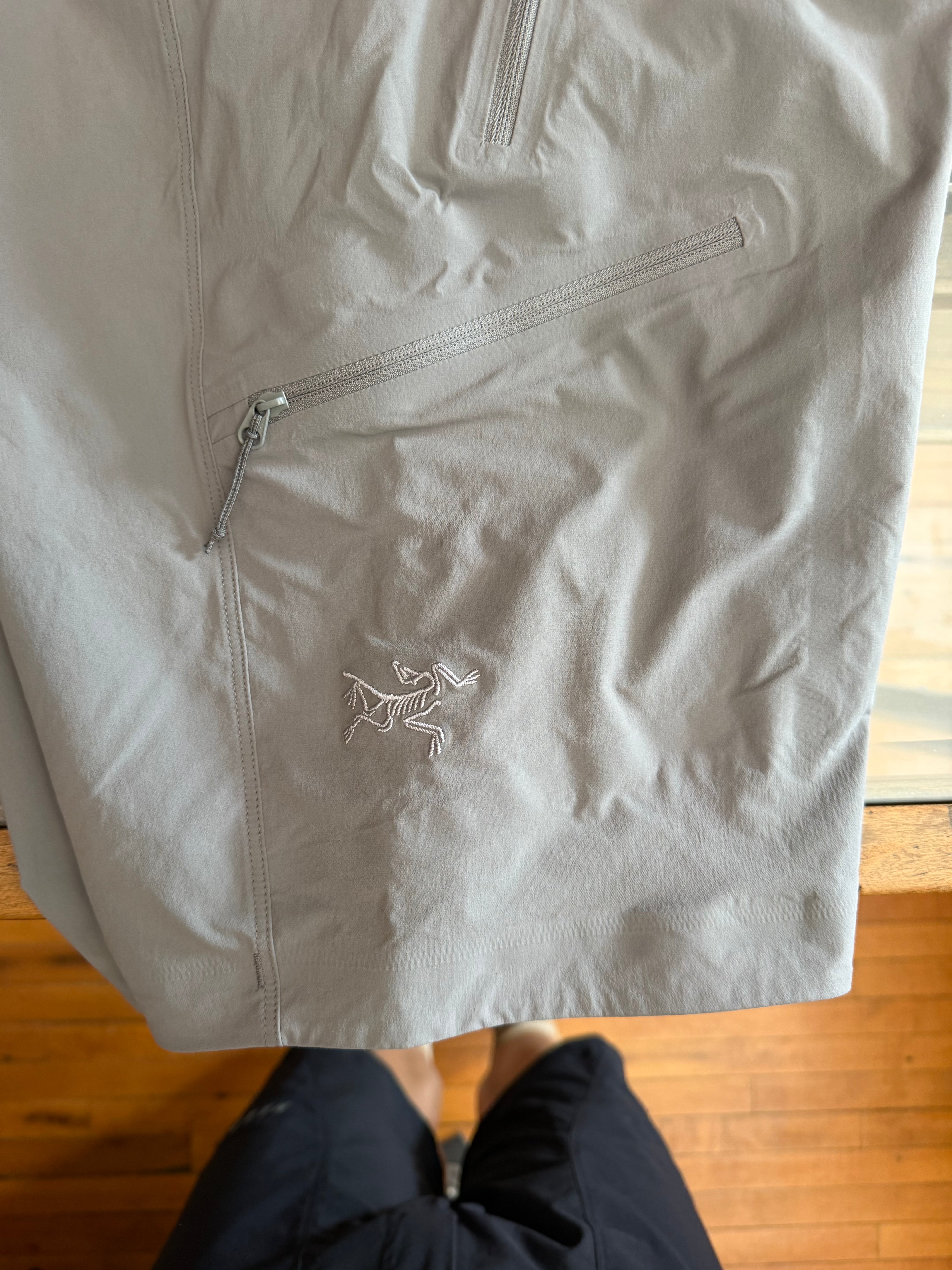 Close-up front view of light gray men's jacket with Arcteryx logo, zipper pocket, and fabric texture details.