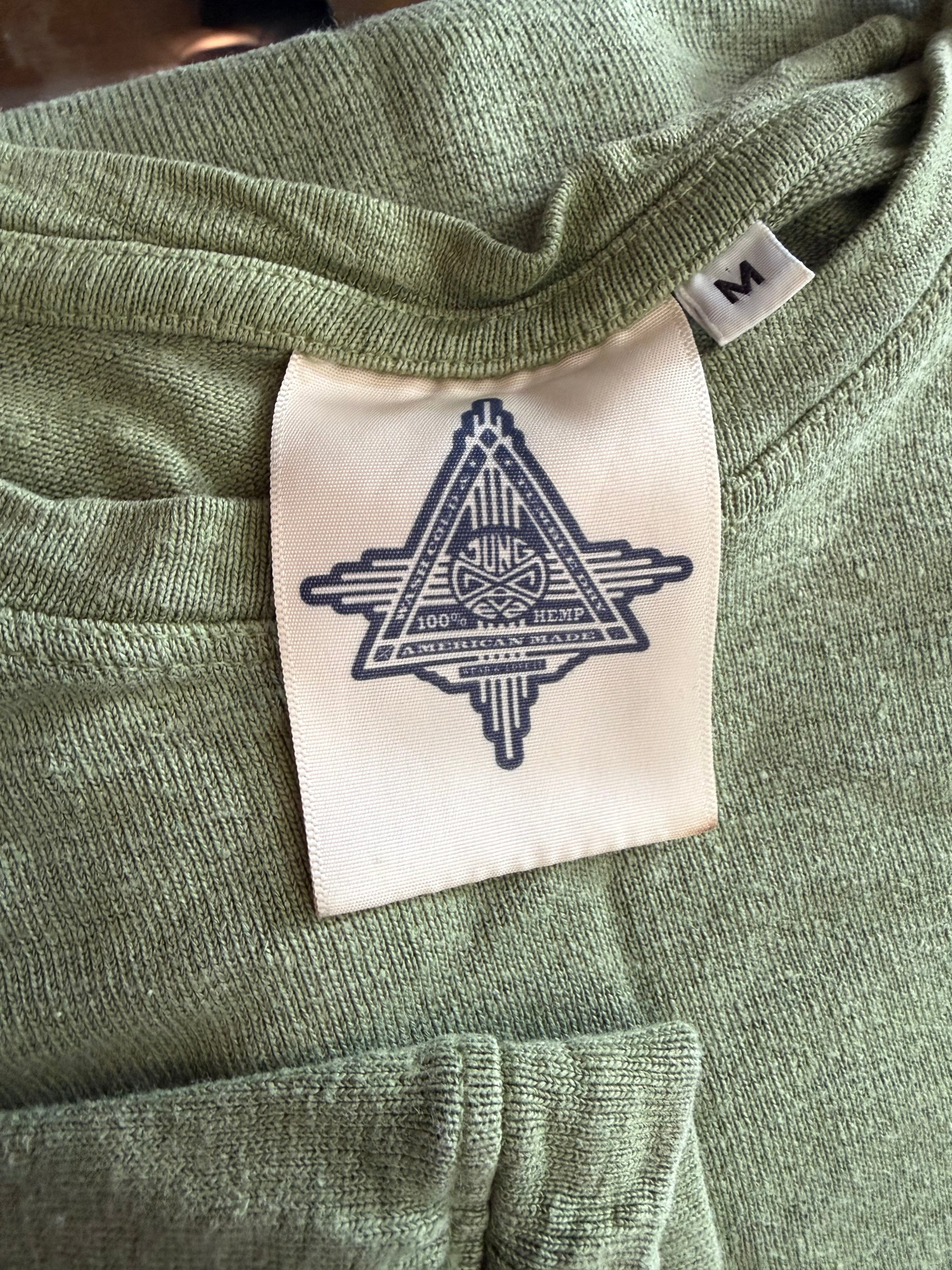 Close-up of sage green knit shirt collar with visible brand tag, fabric texture, and size M label, showing the inside neck detail.