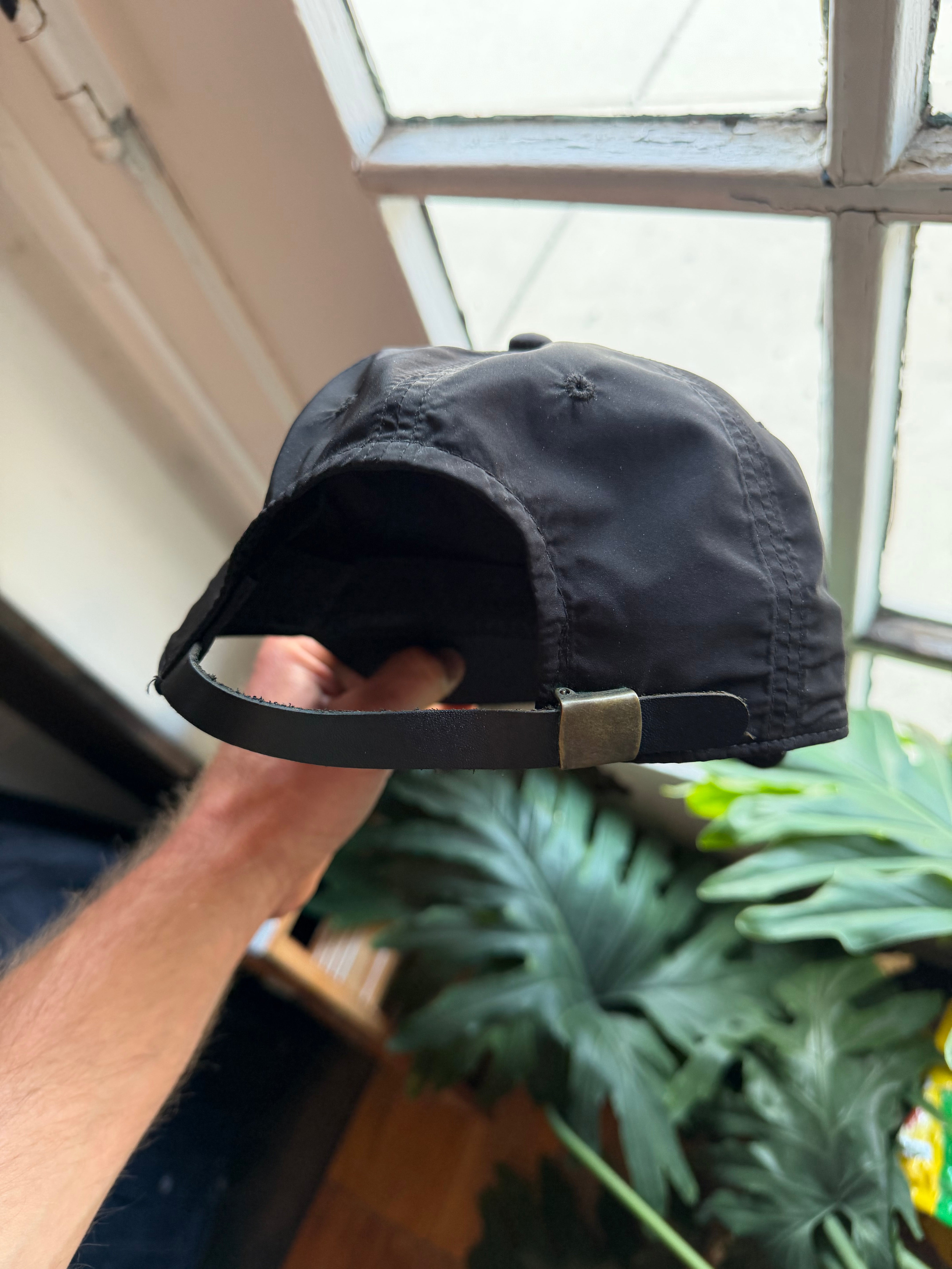 Back view of black cap with adjustable strap, held by hand near window with green plant background