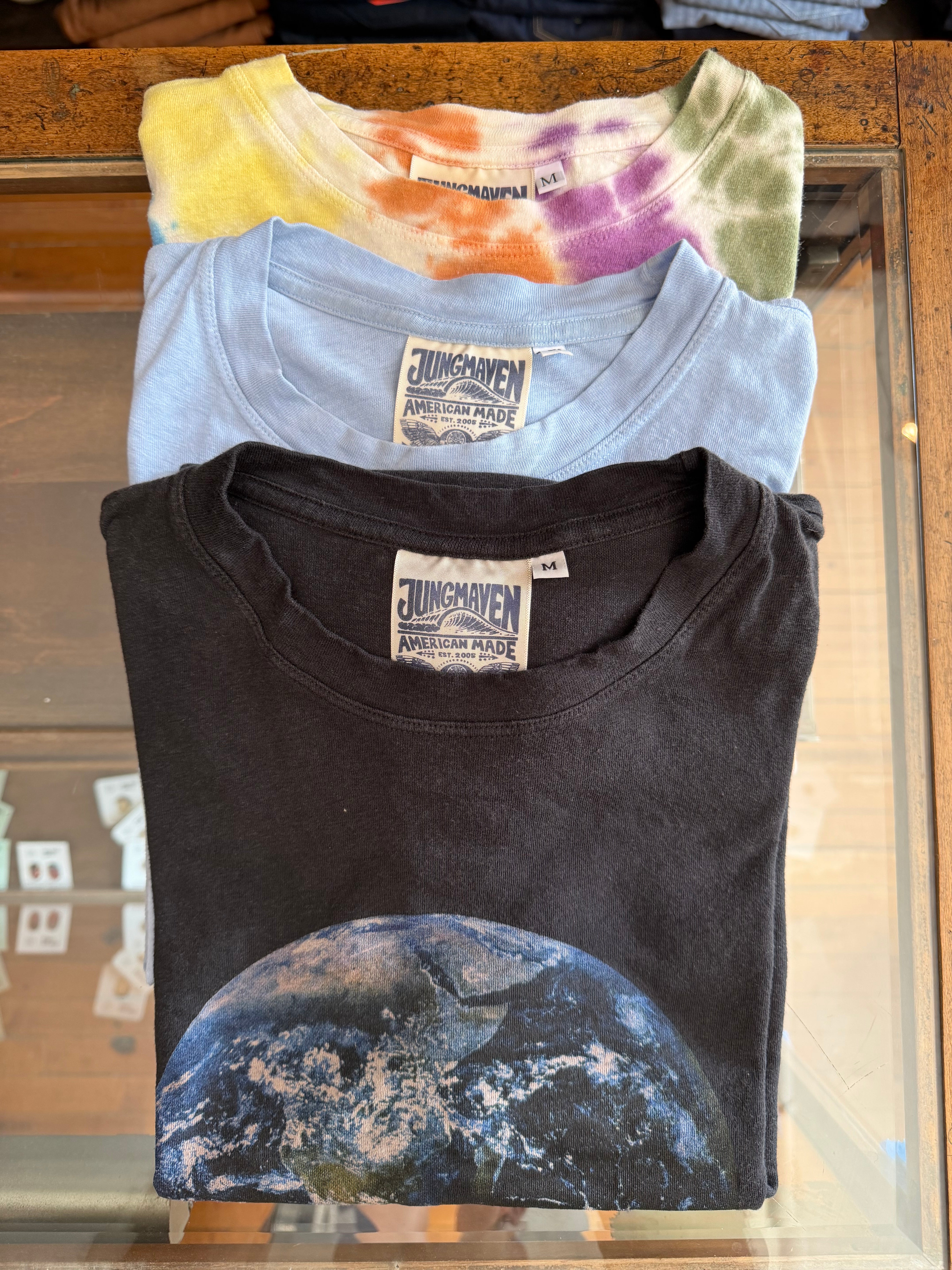 Front view of folded Jungmaven tees: black Earth print, light blue, and tie-dye. American-made, size M tags visible.