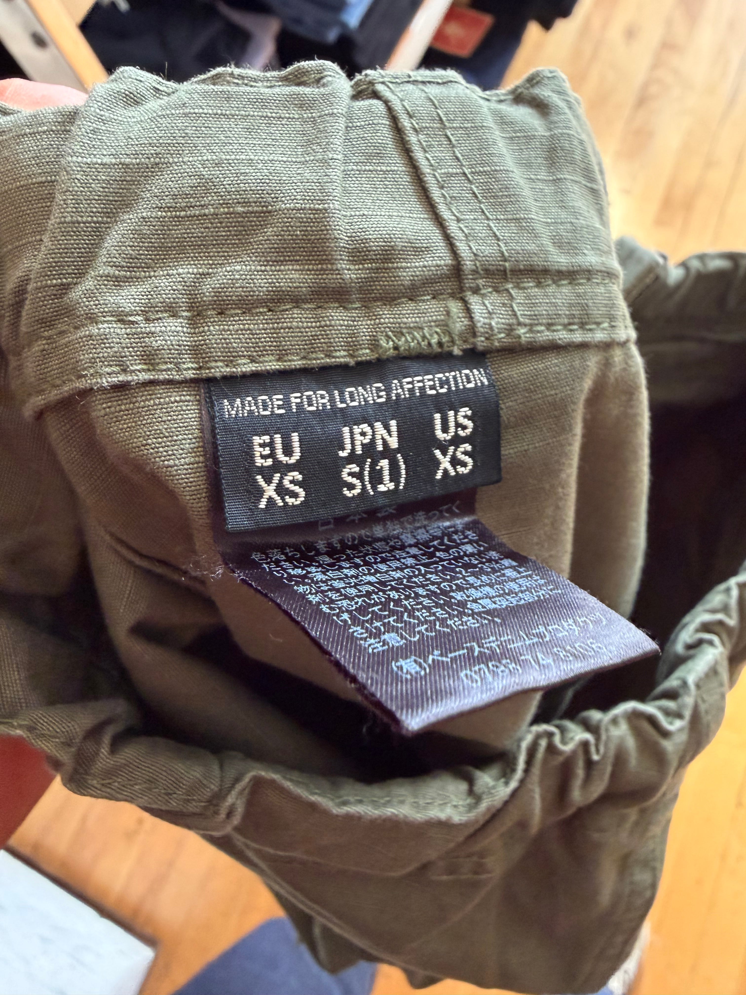 Close-up of olive green pants waistband inner back tag, showing size and care labels, Dant apparel.