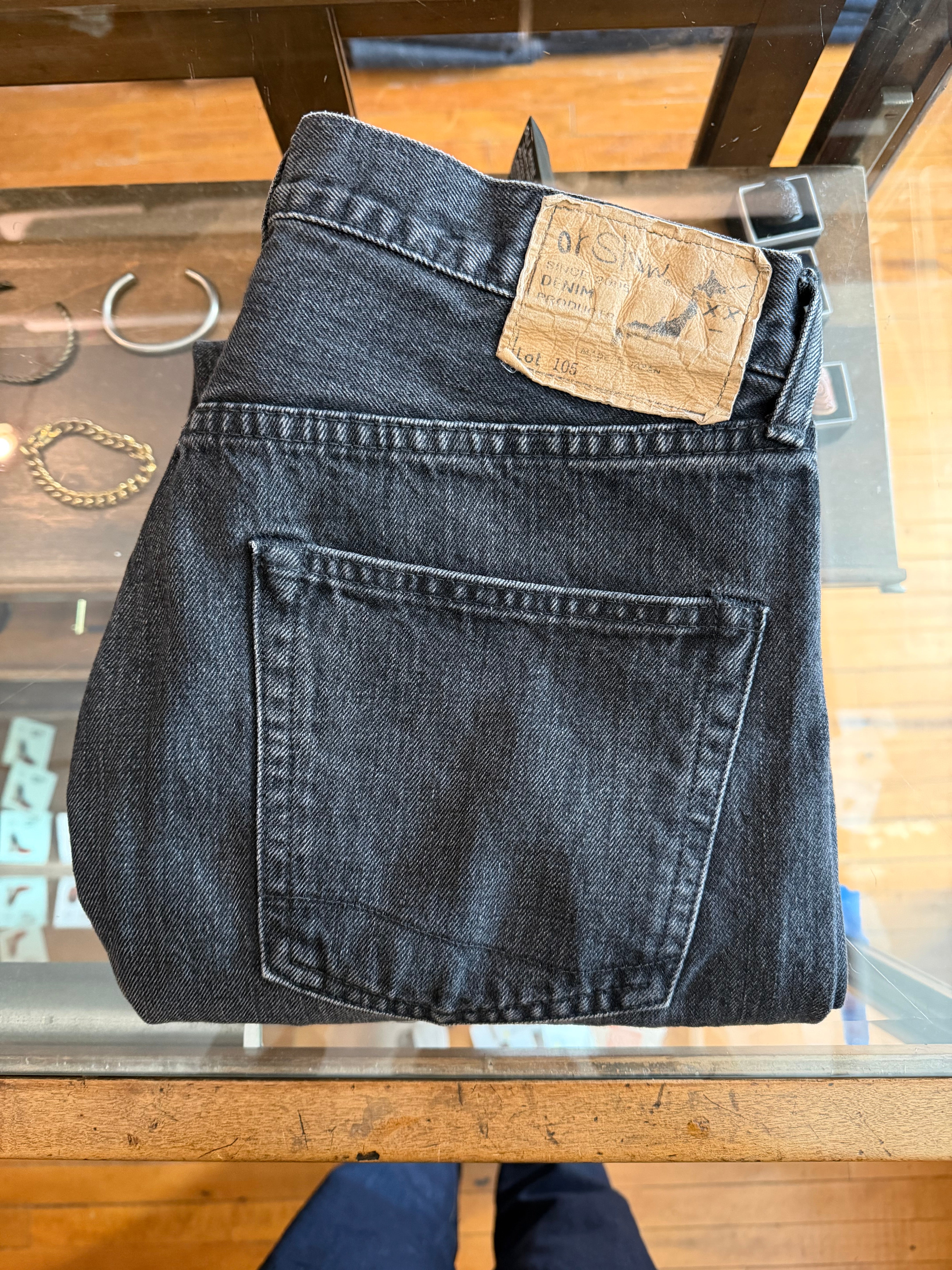 Close-up of folded black selvedge denim jeans back side, visible logo patch, displayed on glass table at Dant, Indianapolis.