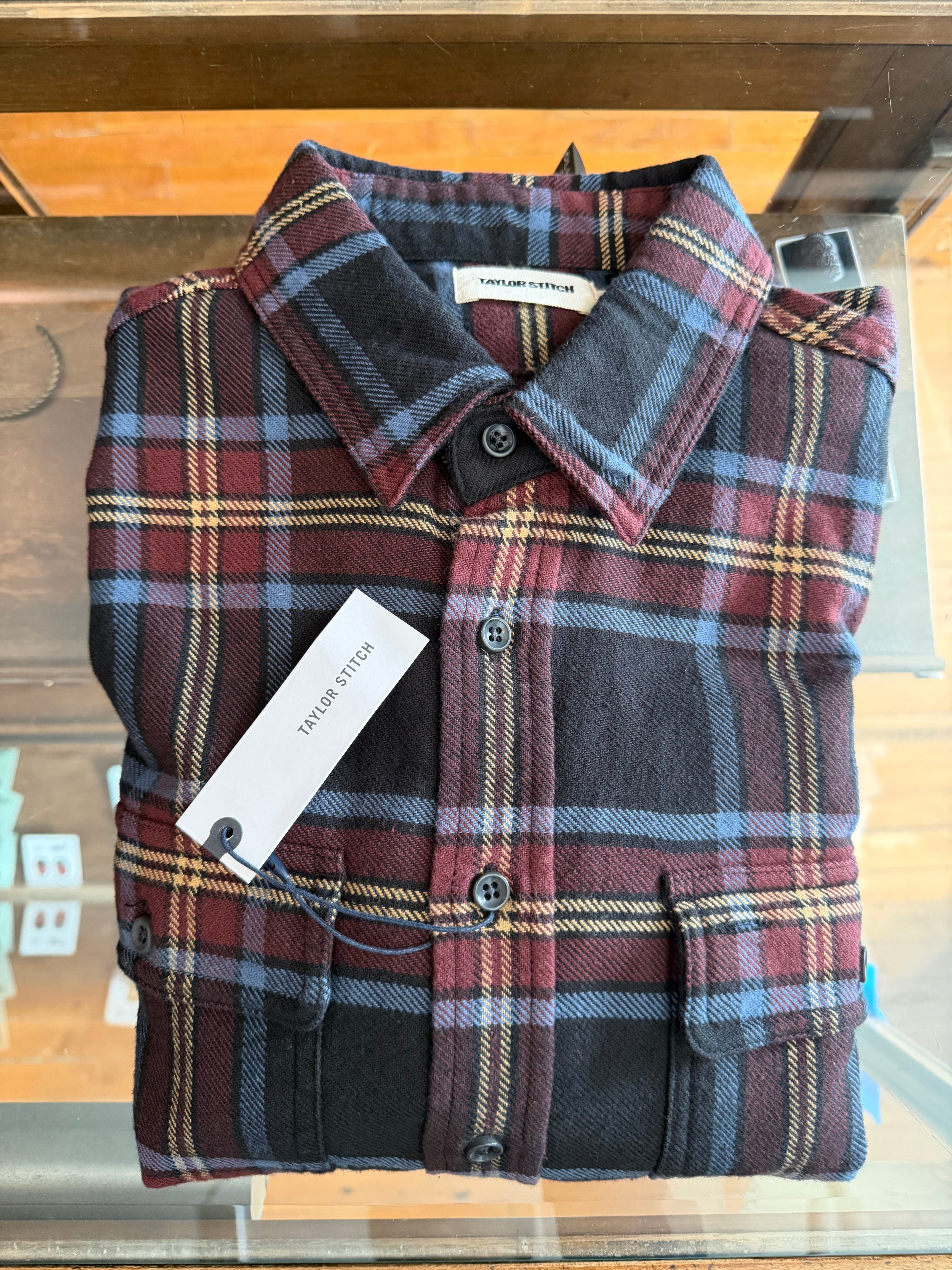 Front view close-up of folded Taylor Stitch men's plaid flannel shirt with collar, buttons, and chest pockets on display at Dant shop.