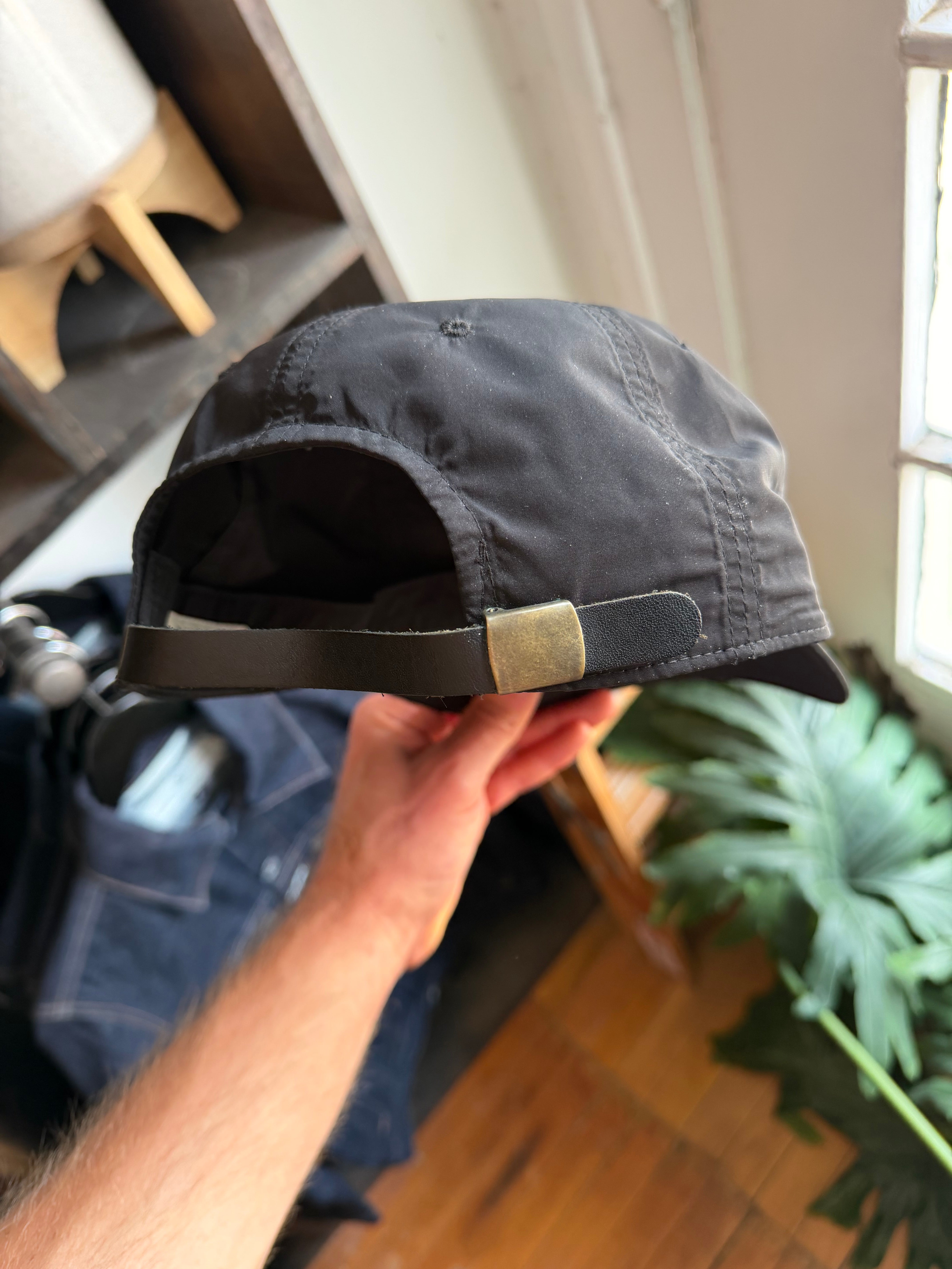 Close-up of black cap's back, showing brass buckle and leather strap, held by hand.