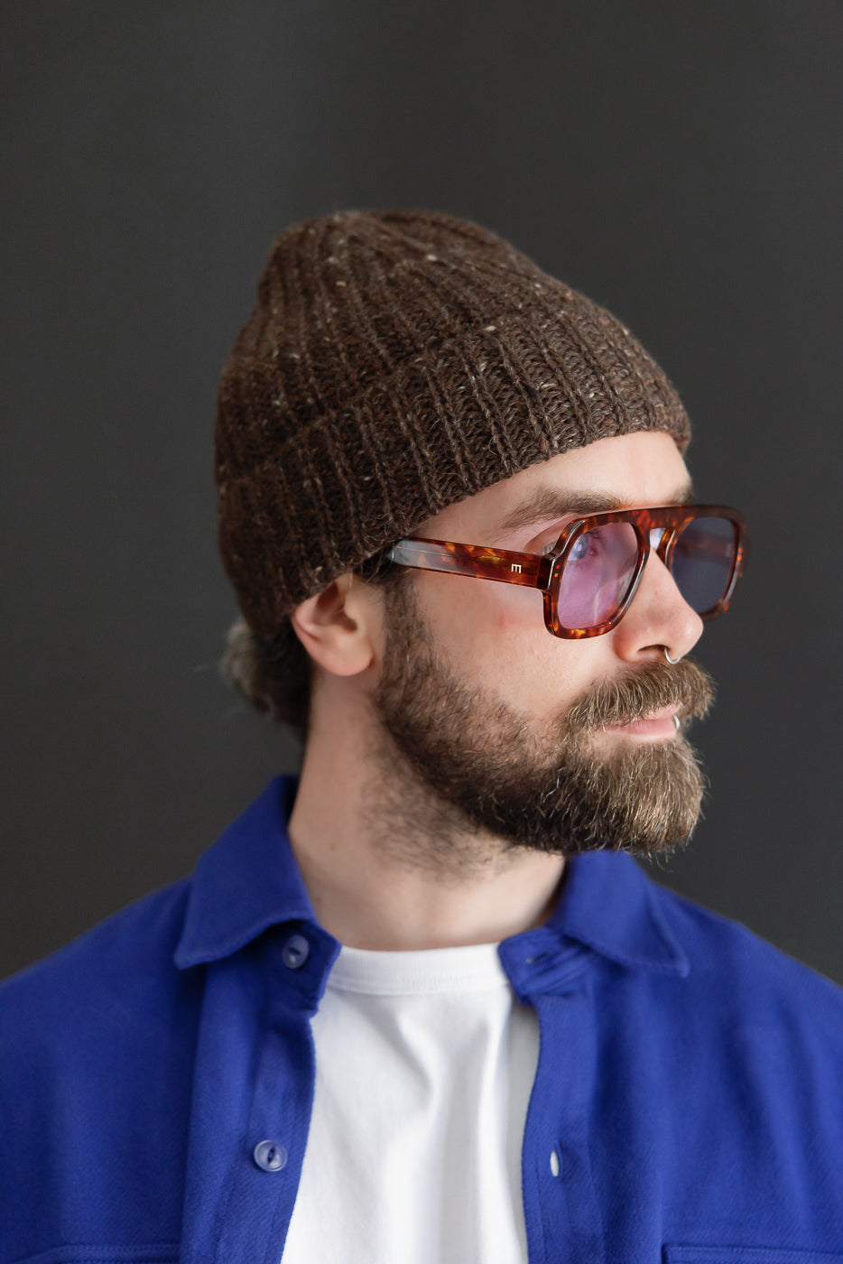 Close-up, side profile of a male model wearing a brown knit beanie and tortoiseshell glasses.