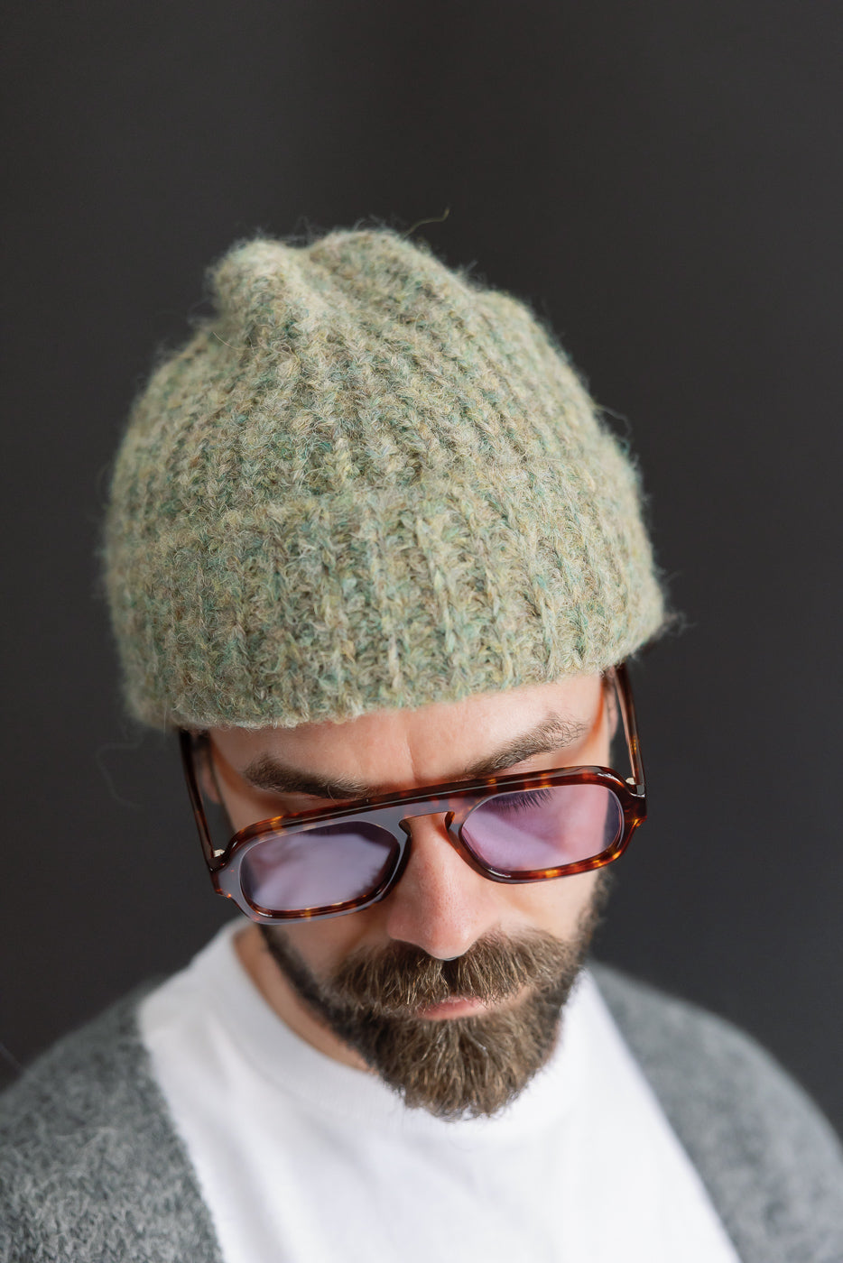 Close-up front view of a male model wearing a textured green knit beanie and sunglasses.