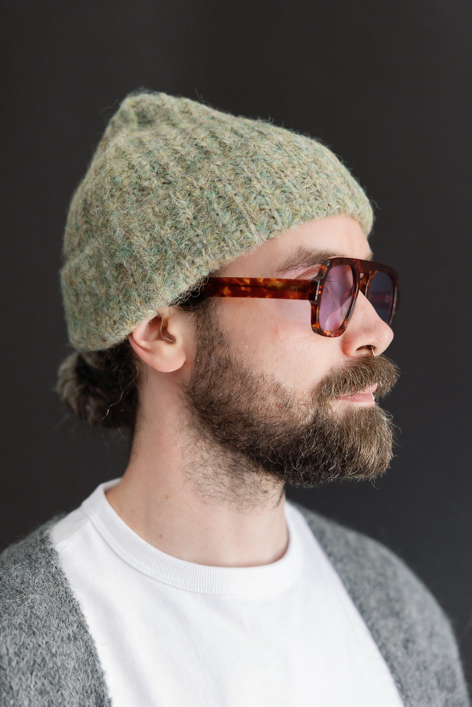 Close-up profile view of a male model wearing a textured green knit beanie and tortoiseshell sunglasses.